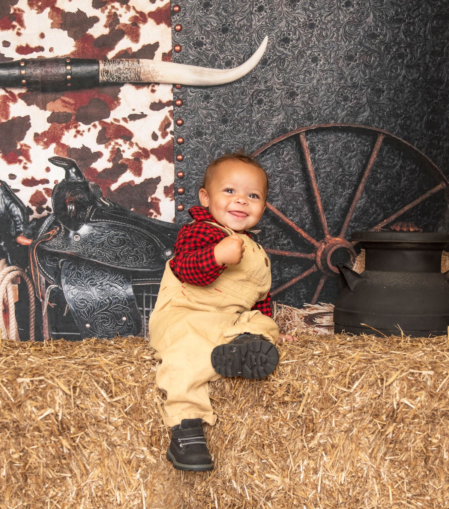 A young child in tan overalls and red shirt sits on hay bales in a rustic western-themed photo setting with wagon wheel decor.