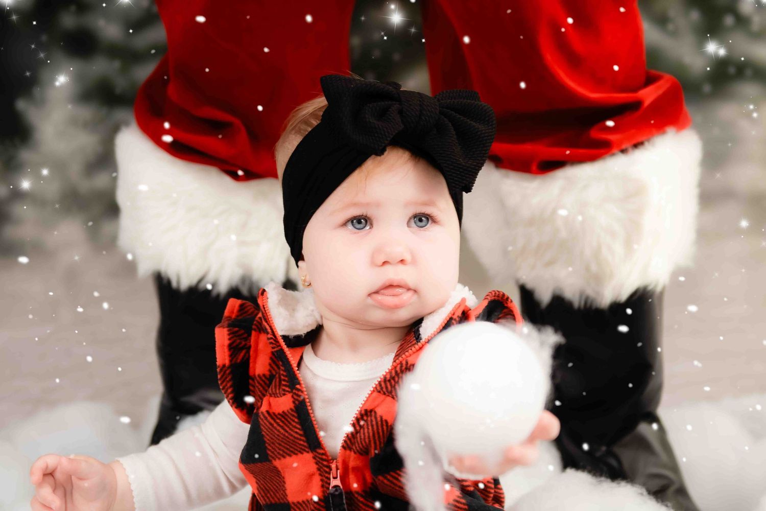 A child in a buffalo plaid outfit and black bow sits with Santa in a snowy winter portrait setting.