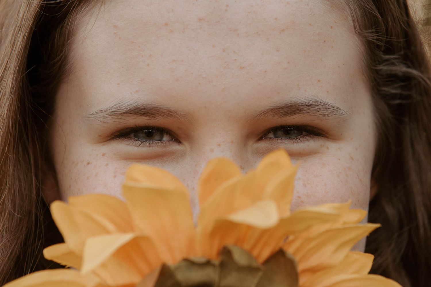 A large yellow sunflower bloom fills the lower portion of the frame while someone peers over it from behind.