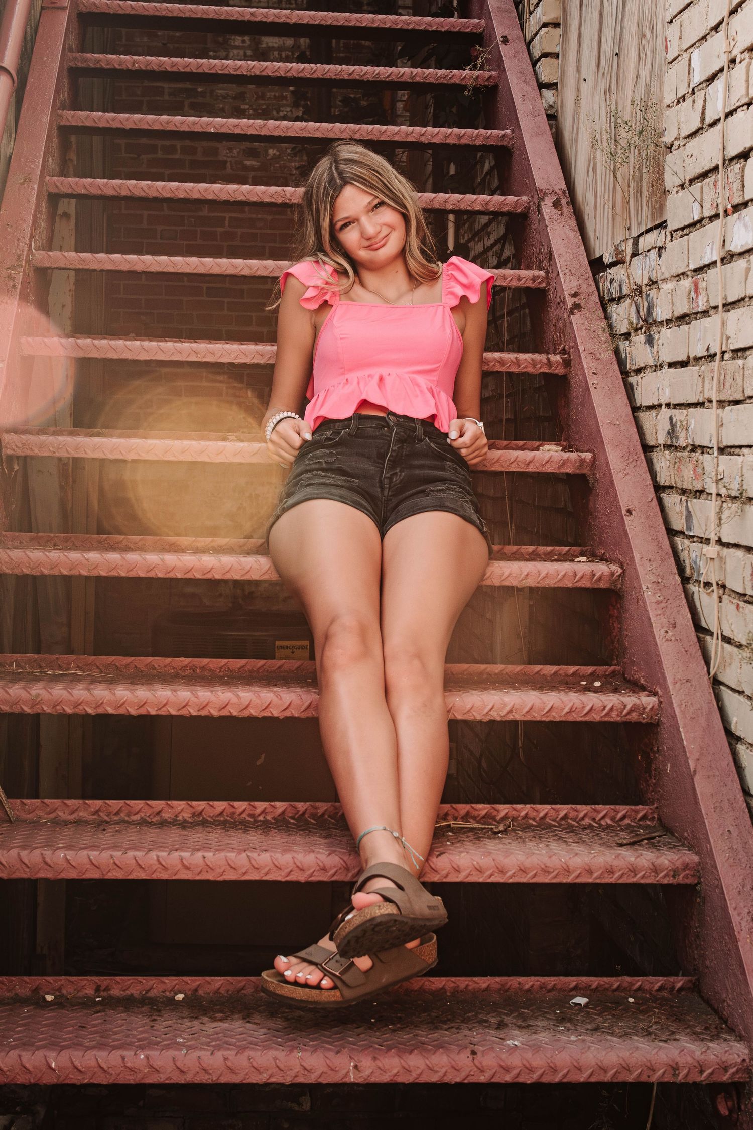 Person wearing a pink top and dark shorts relaxing on rustic red metal stairs outside a brick building.