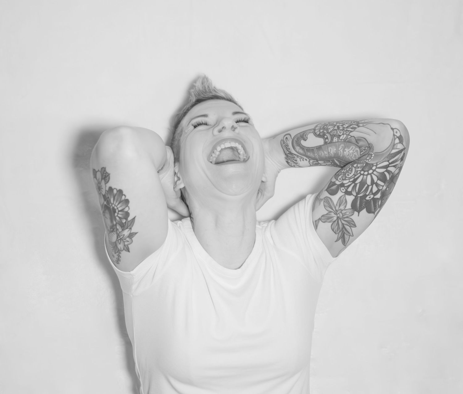 Person in white t-shirt throws their head back laughing, showing off intricate arm tattoos in black and white photo.