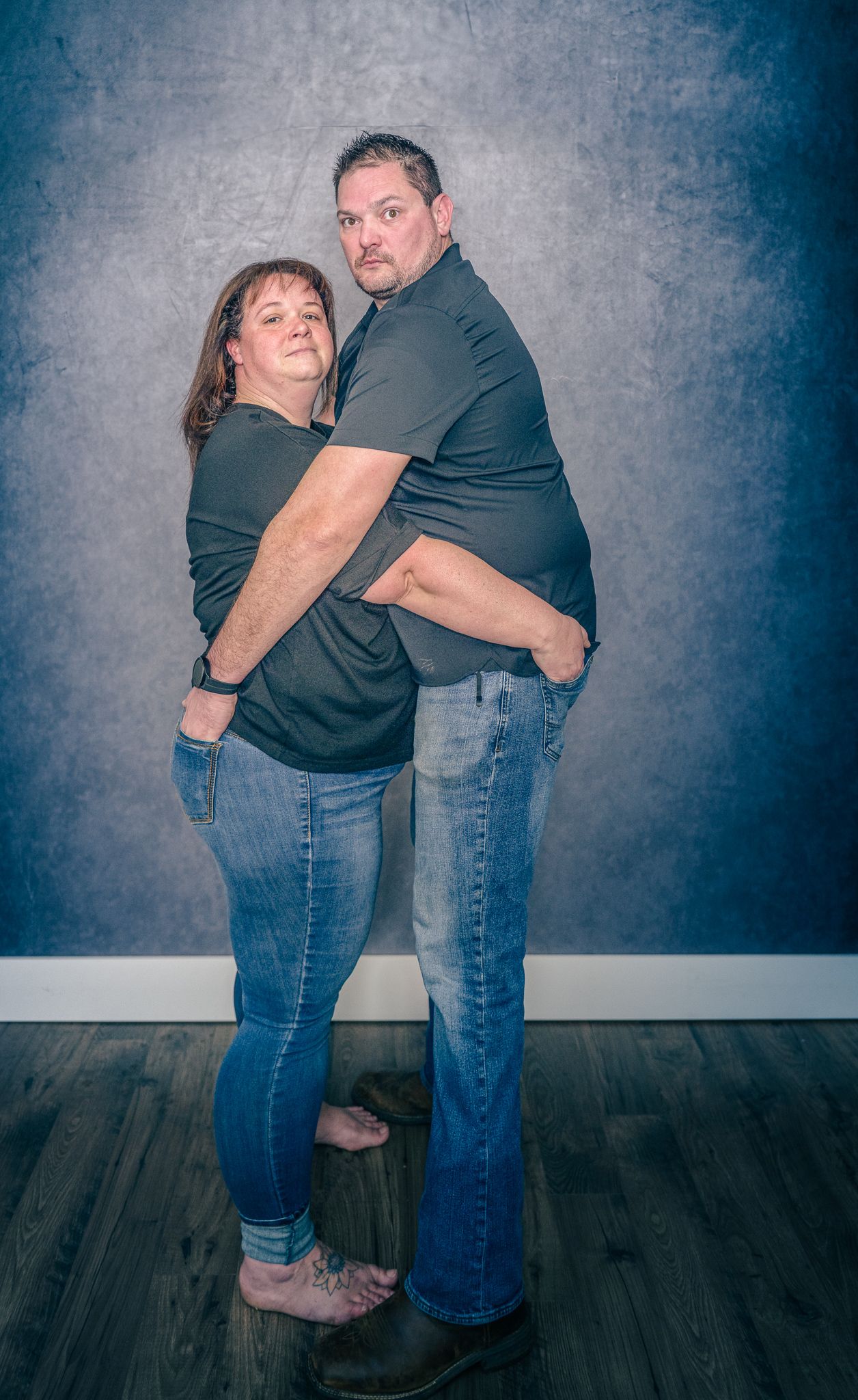 A couple in matching dark shirts and jeans pose together for a playful studio portrait against a blue-gray backdrop.