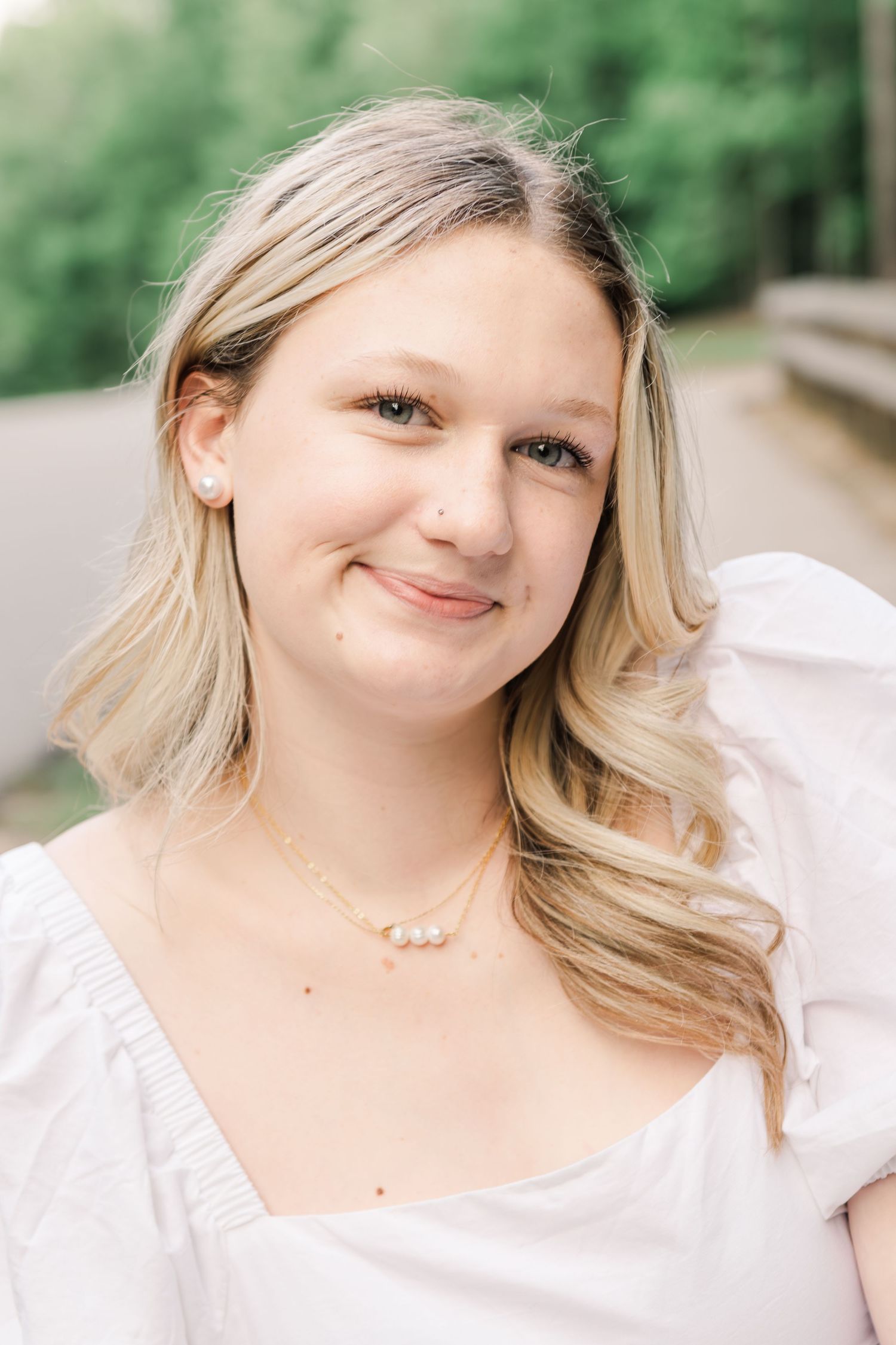 Portrait outdoors with natural lighting showcasing a delicate pearl necklace and white blouse against a blurred green backdrop.