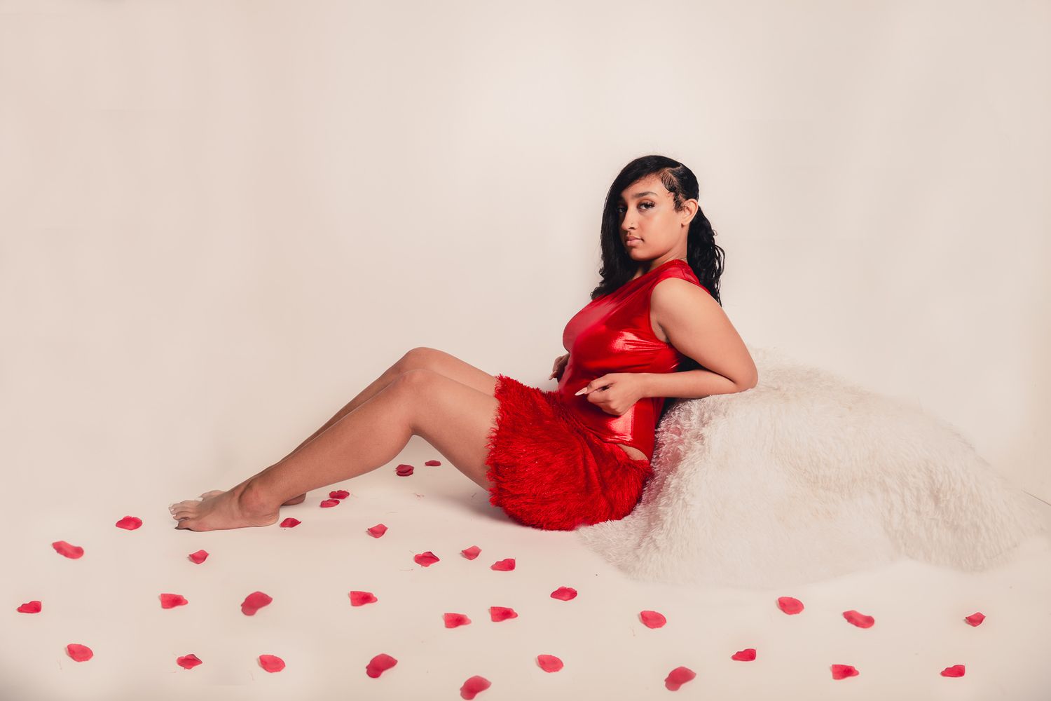 Figure reclining on white fabric in red dress with scattered rose petals for romantic valentines photoshoot.