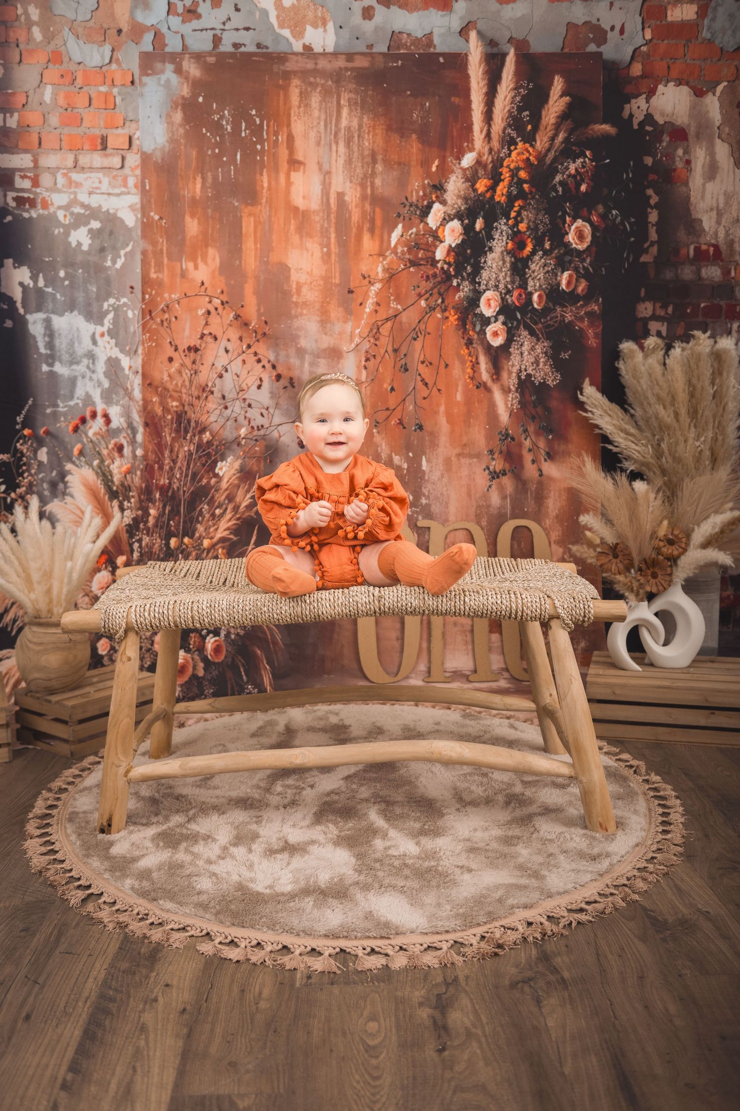 A child in orange clothing poses on a wooden bench with a rustic fall-themed background and floral decorations.