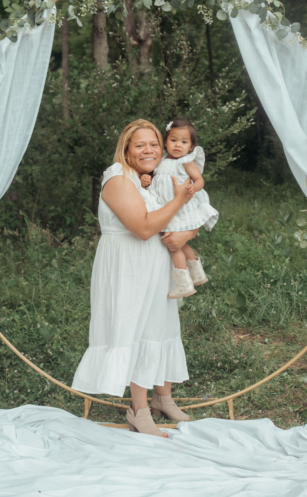 A white floral arch frames an outdoor photo shoot with two people in matching white dresses on a fabric backdrop.