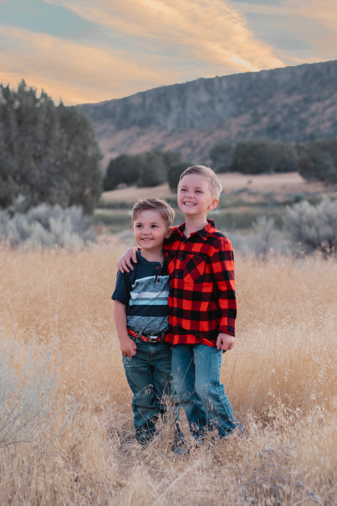 Two children stand together in a golden field of tall grass with mountains and a sunset sky in the background.