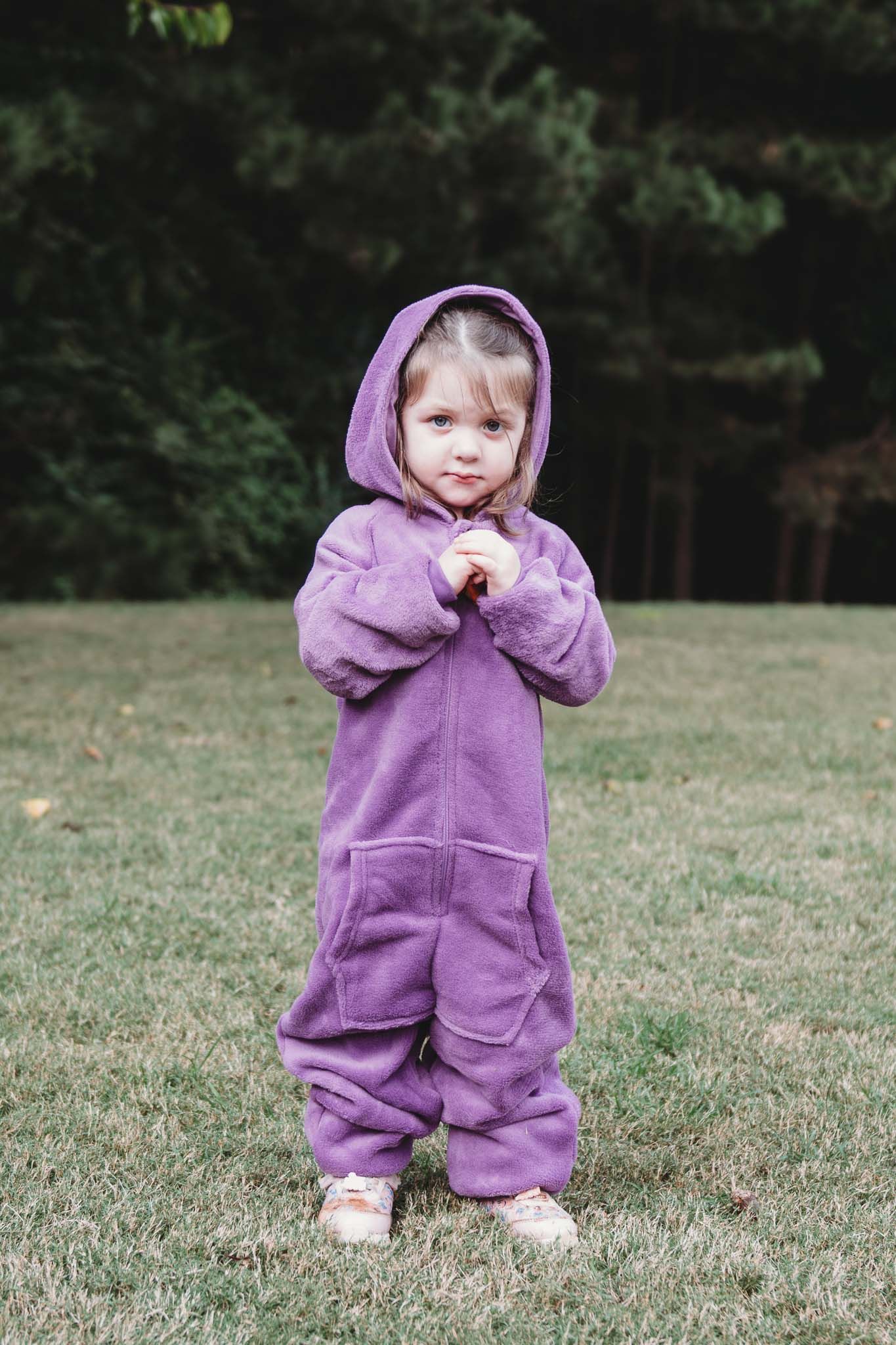 Small child wearing a cozy purple hooded jumpsuit stands on grass with trees in the background.