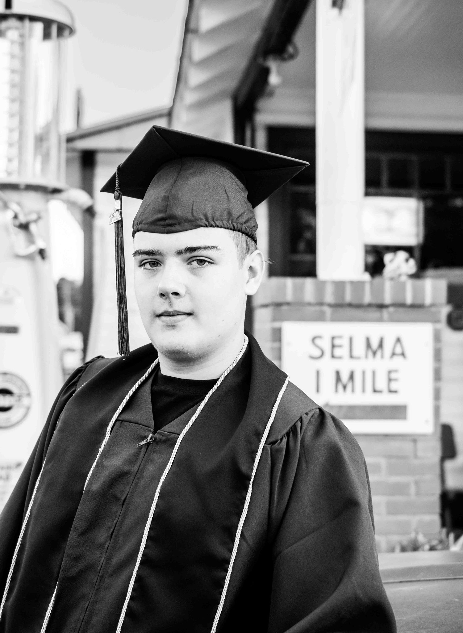 Graduate in black cap and gown poses in front of vintage Selma 1 Mile sign in black and white photo.