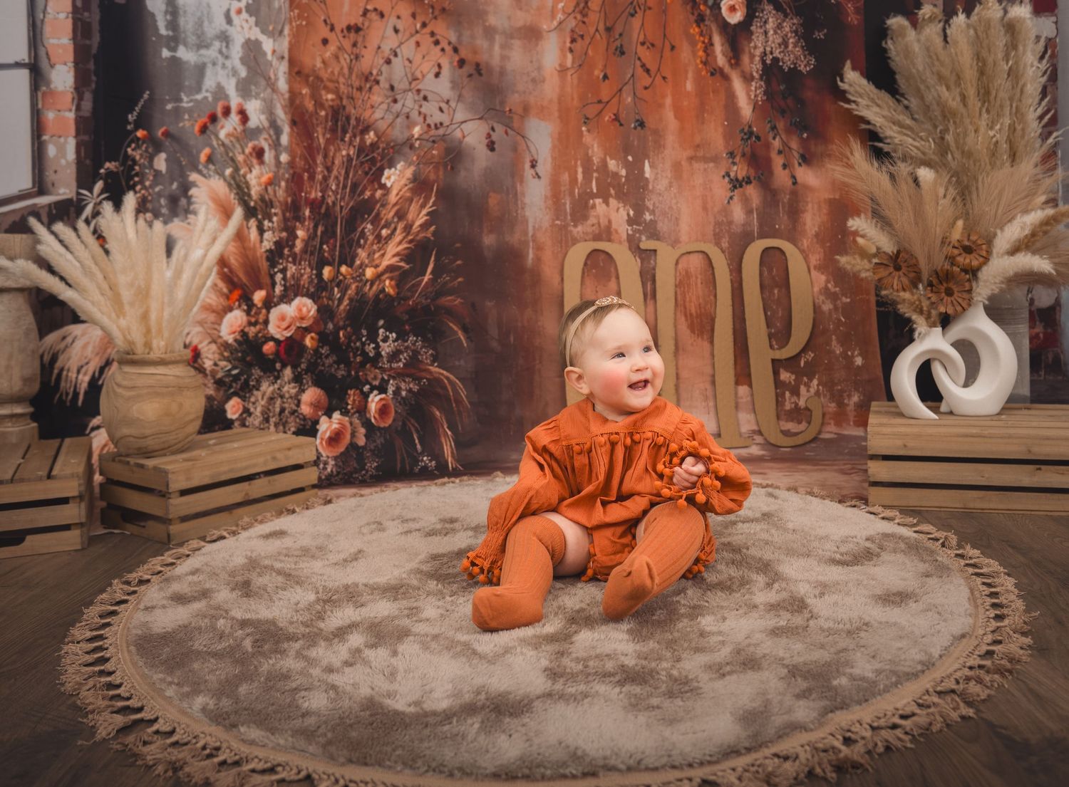 A small figure in bright orange sits on a circular rug surrounded by rustic fall decor and dried floral arrangements.