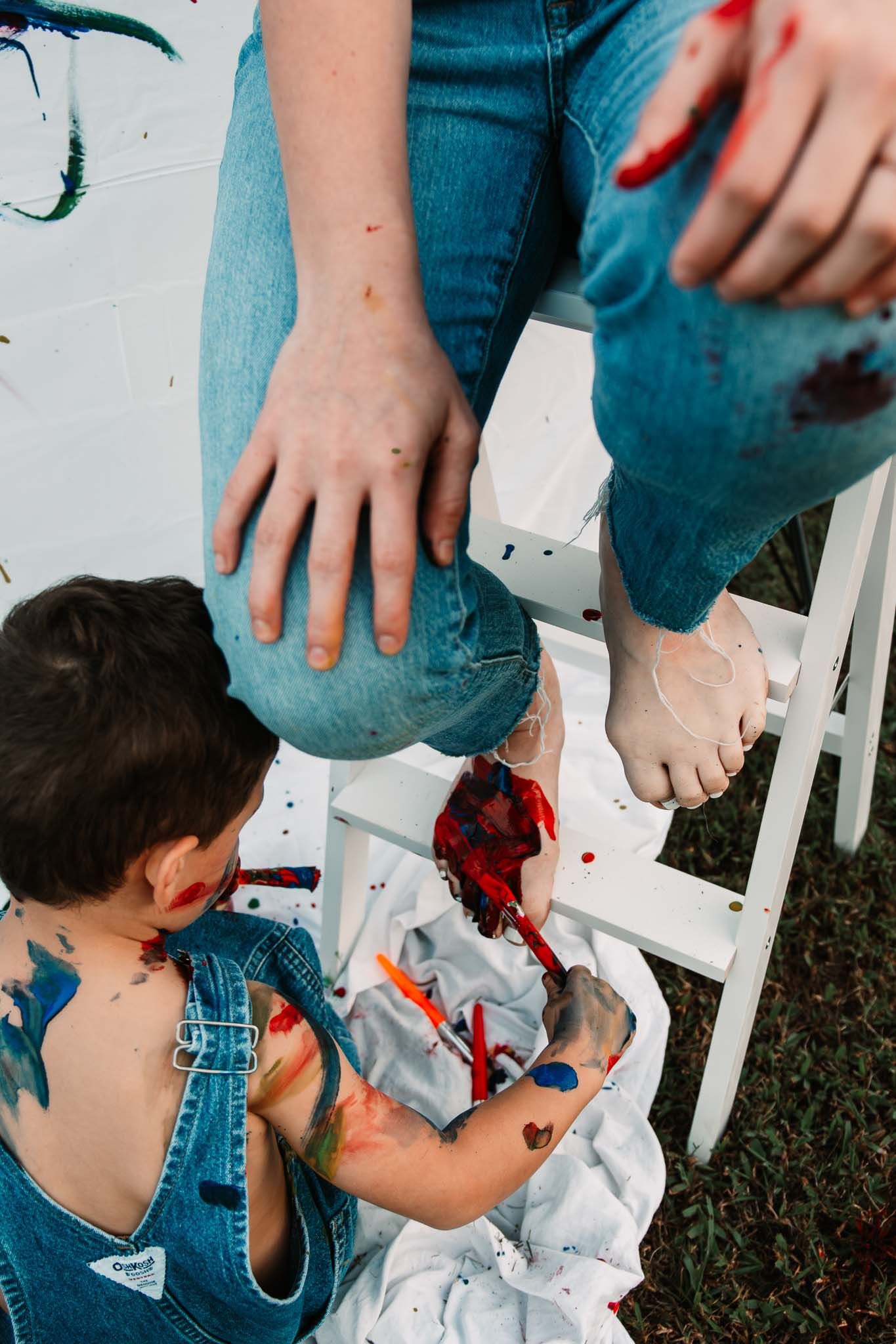 Small child in overalls getting messy with red paint while playing outside by a white ladder.