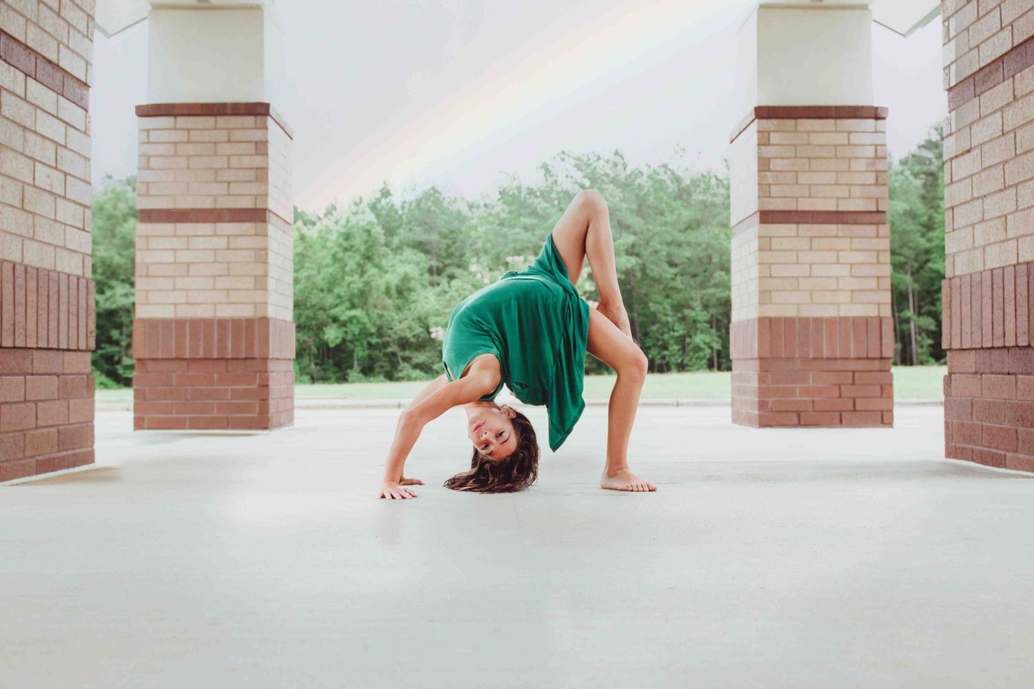 A dancer in a green leotard performs a graceful backbend pose between brick columns on a concrete walkway.