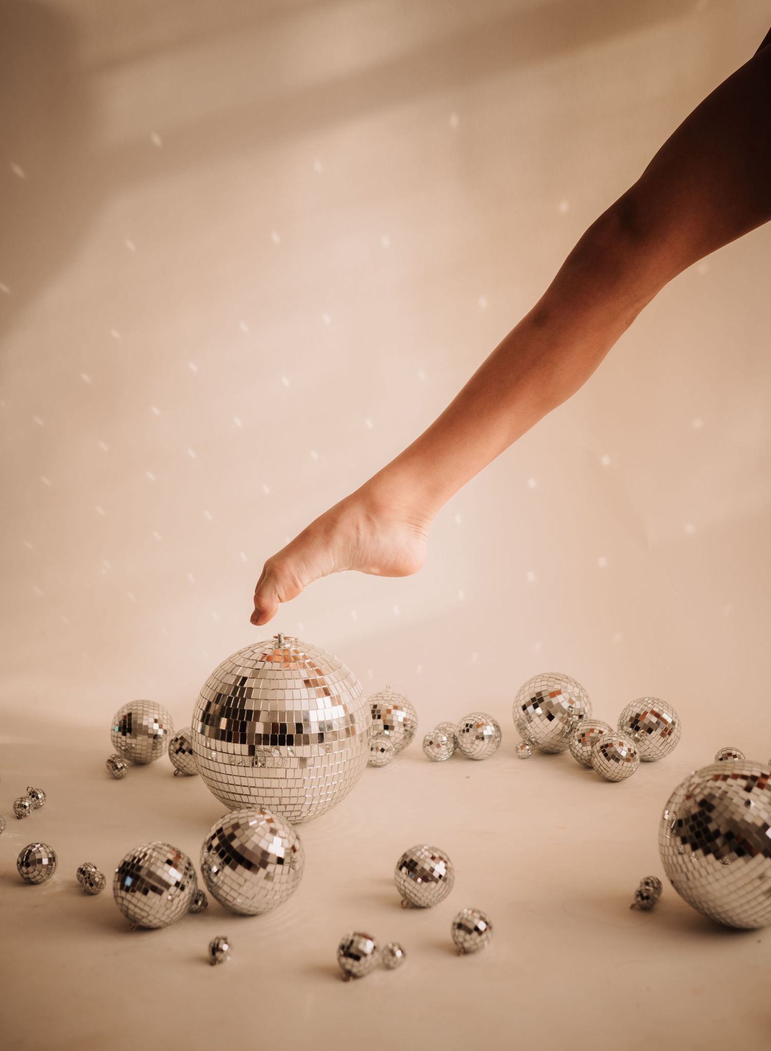 A bare foot reaches toward scattered silver disco balls of varying sizes against a warm beige background.