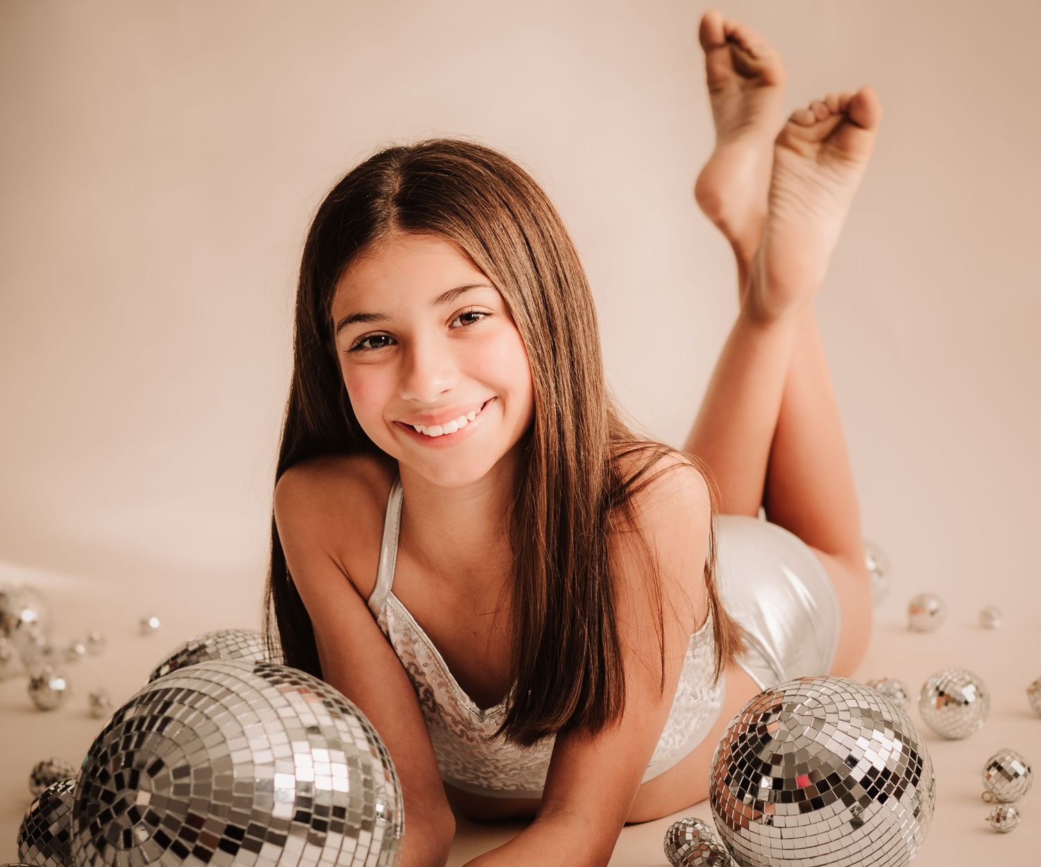 Portrait of someone posing on a floor with two disco balls against a beige background.
