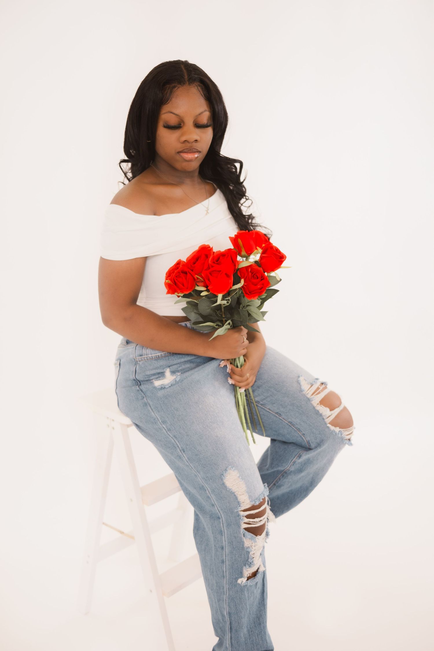 Person in distressed jeans and white off-shoulder top poses with red roses against white backdrop in artistic photoshoot.