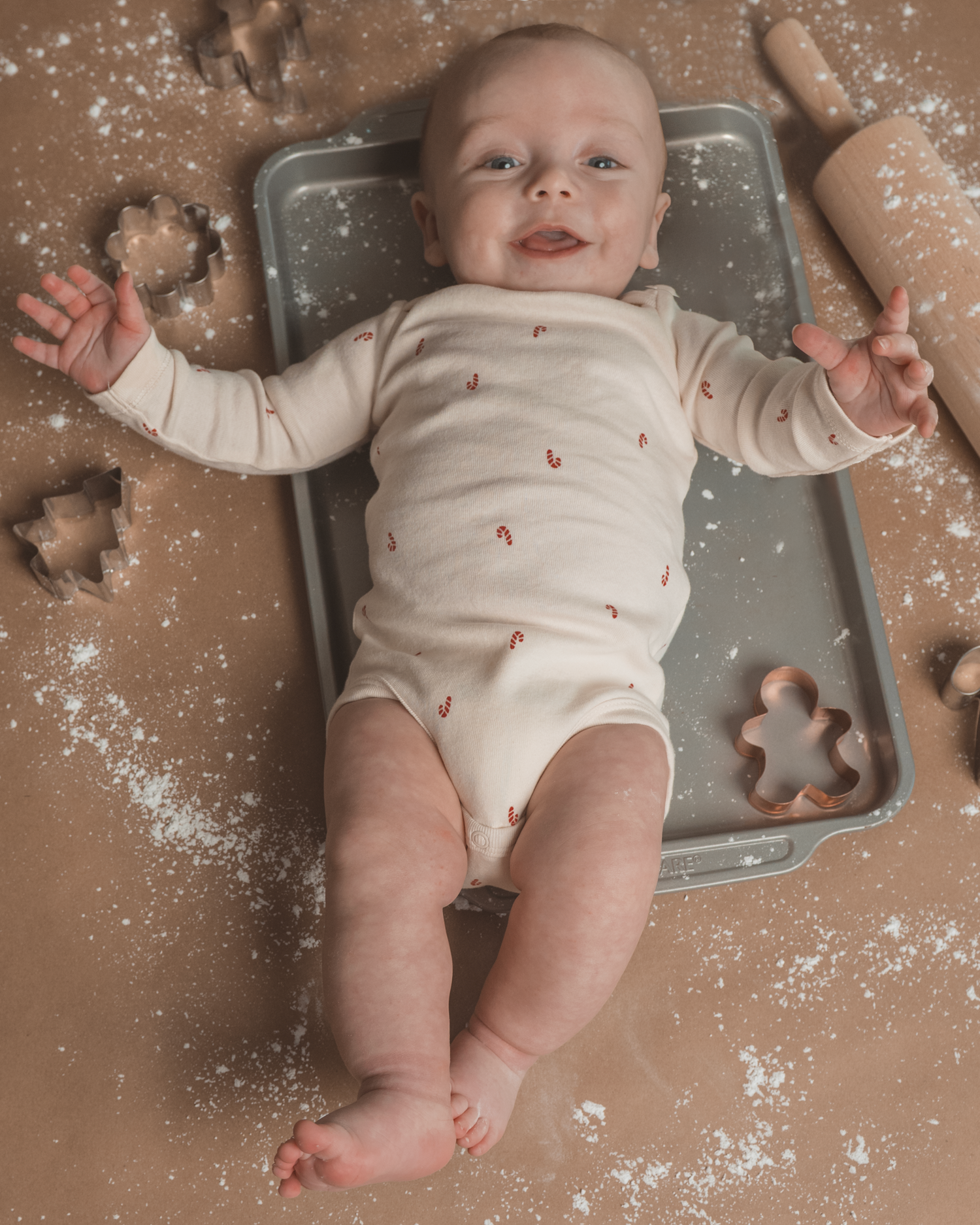 Baby lying on kitchen counter surrounded by flour and gingerbread cookie cutters in festive holiday baking scene.