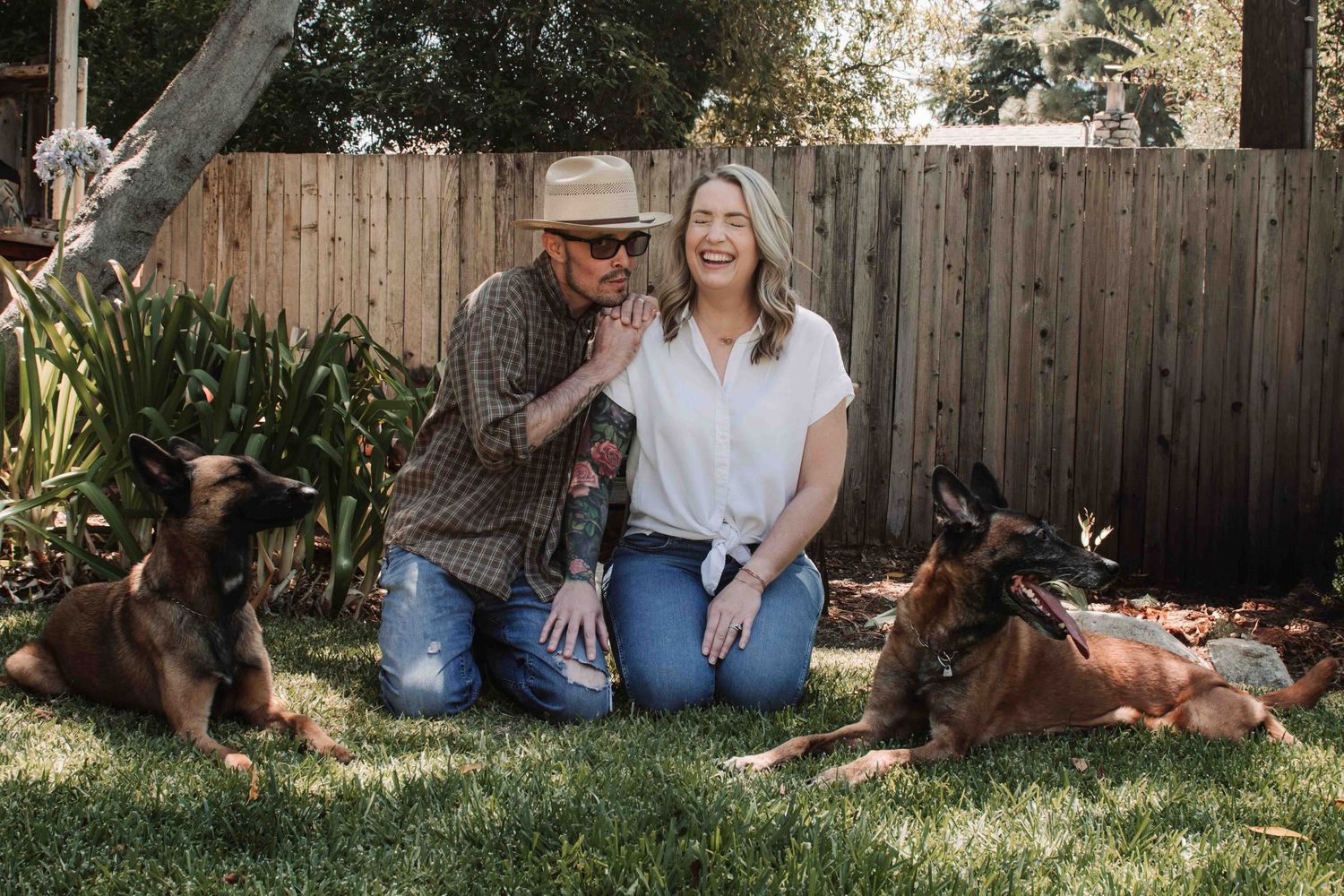 A couple sits together on grass with their brown dog in a backyard setting during golden hour.