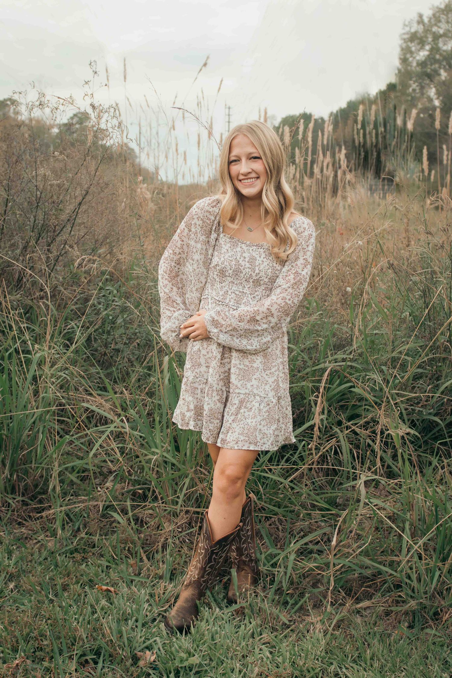 A person in a floral print dress and brown boots poses in a field of tall grass on an autumn afternoon in Smithfield, NC