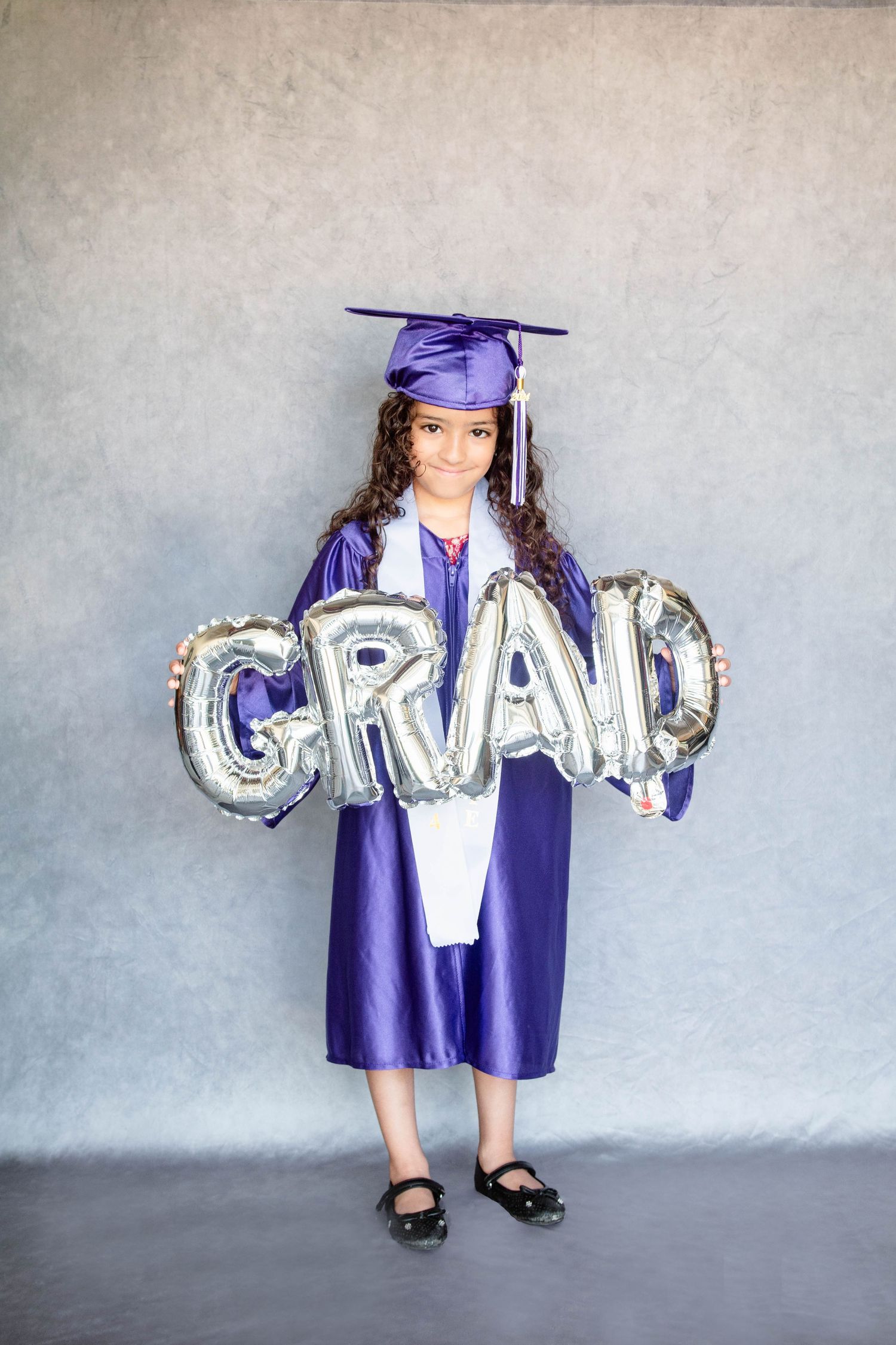 Graduate in purple cap and gown holding silver GRAD balloons stands against gray background.
