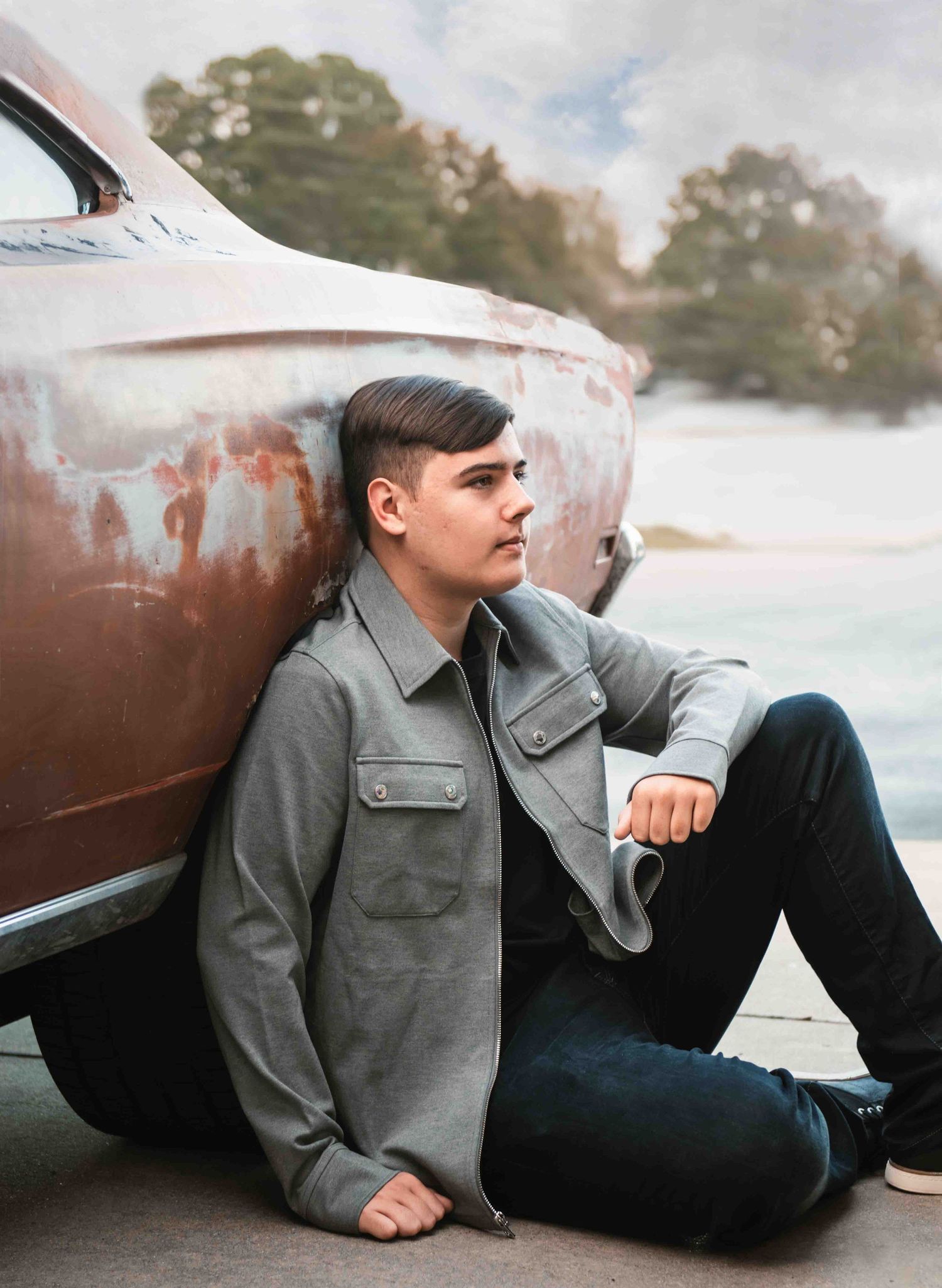 Person in gray utility jacket sits against rustic vintage car exterior in artistic portrait composition.