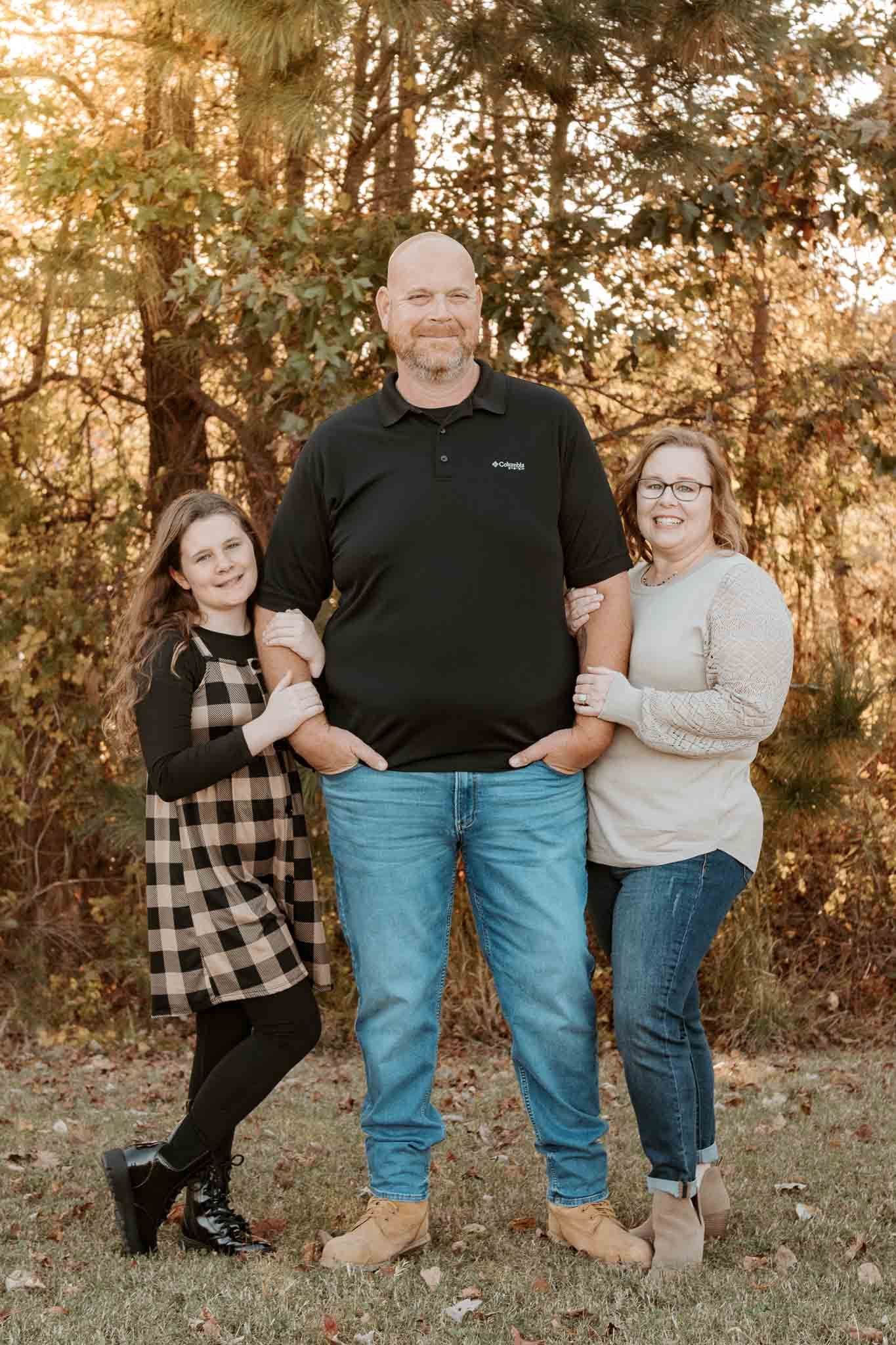 A family of three poses together outdoors with sunlight filtering through autumn trees in the background.