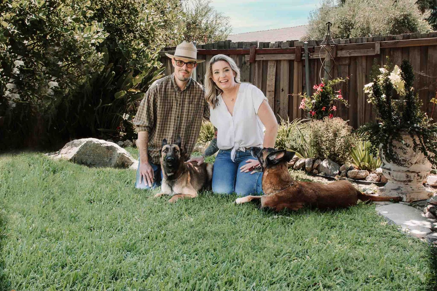Two people sit in a backyard garden with dogs on green grass near a wooden fence and flowering plants.