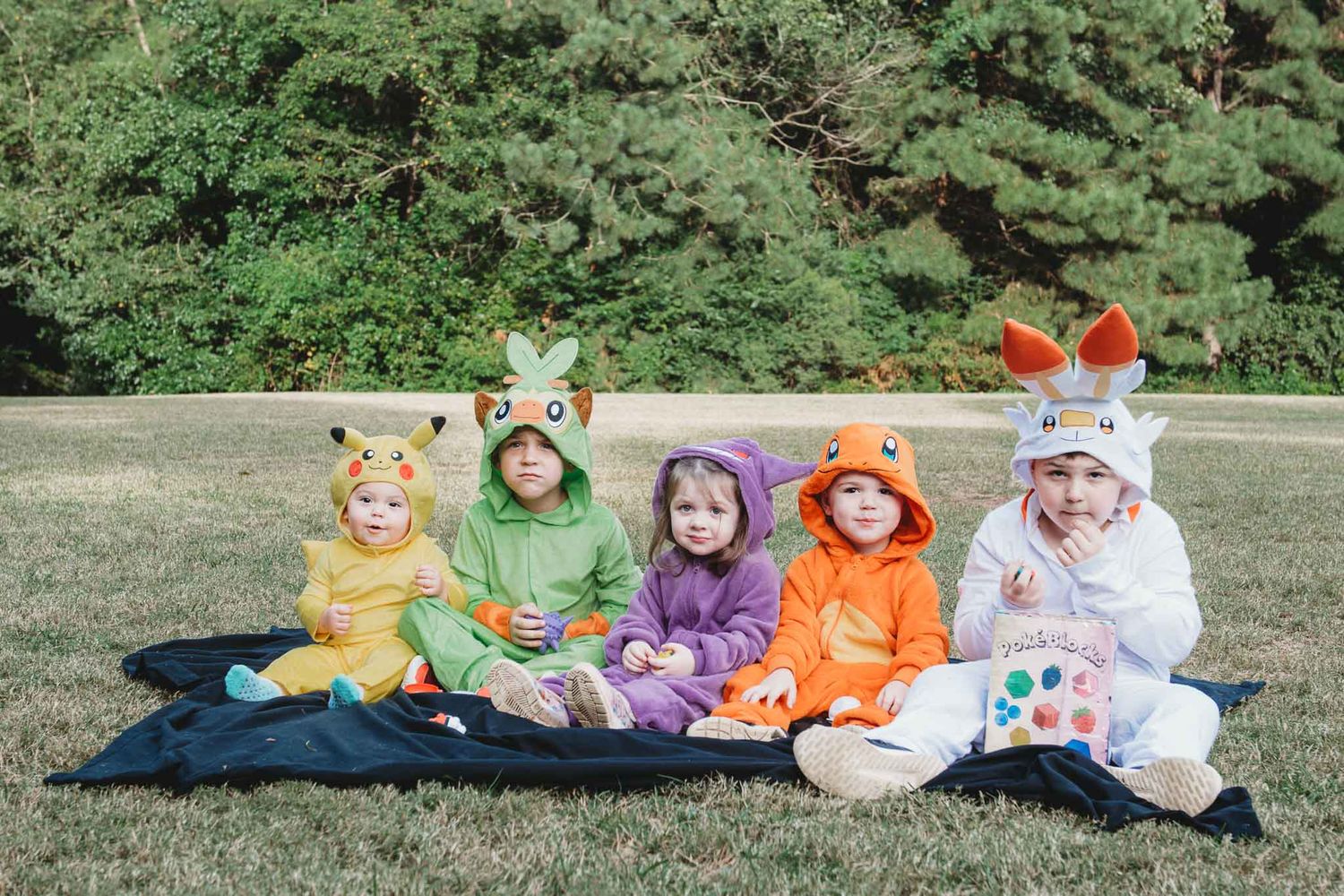 Five people wearing colorful animal onesie costumes sit together on a blanket outdoors on green grass.