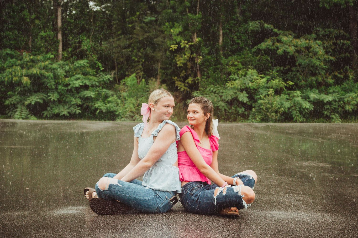 Two people sitting back to back on pavement wearing casual clothing, laughing and sharing a joyful moment together.