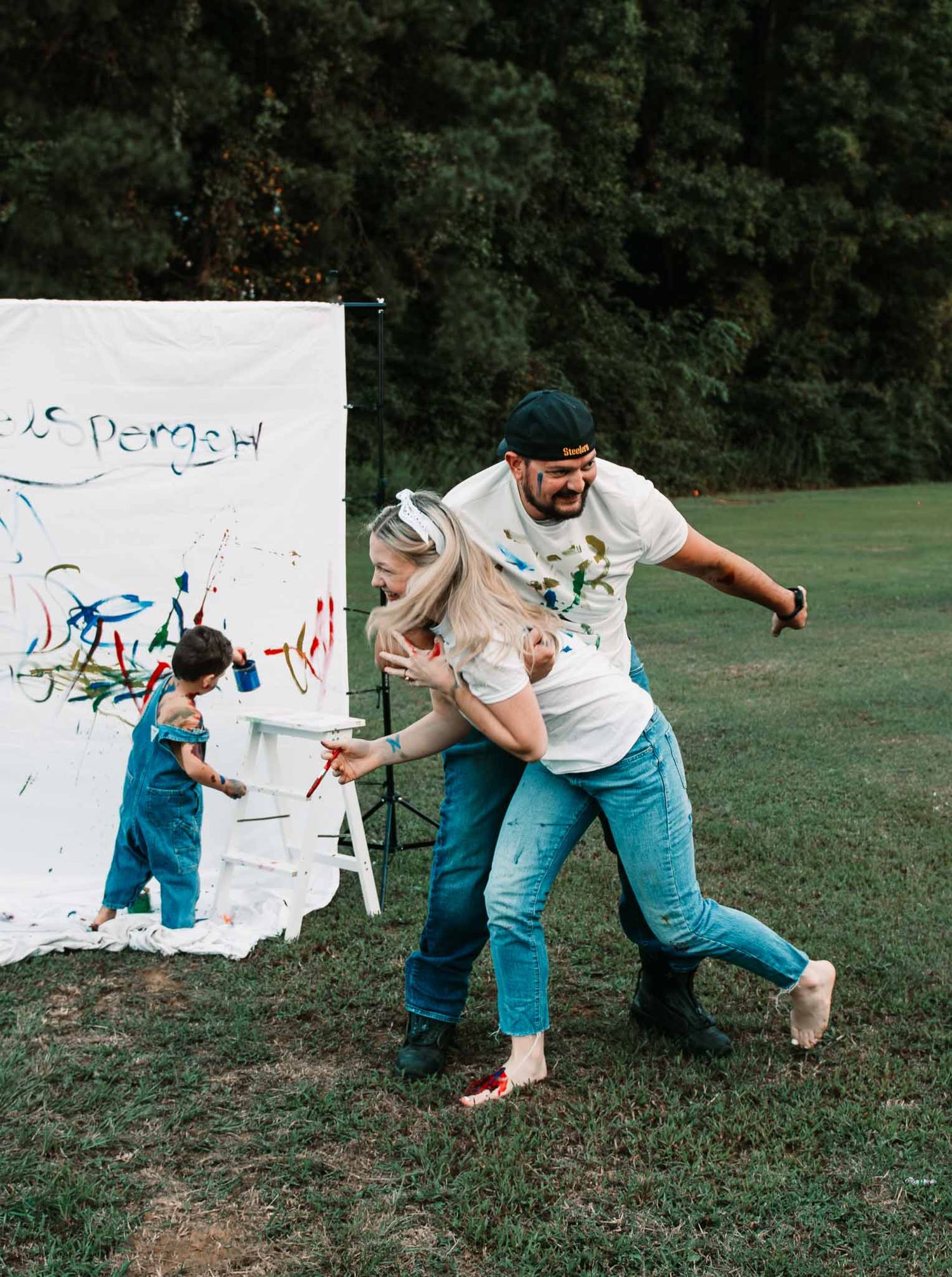 Family creates fun memories with paint by throwing color at a white canvas backdrop outdoors on a grassy area.