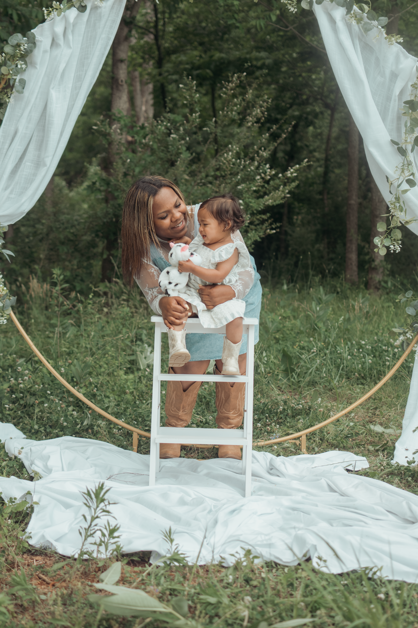 A tender outdoor portrait taken under flowing white curtains and an arch with a ladder used as a prop in a wooded setting.