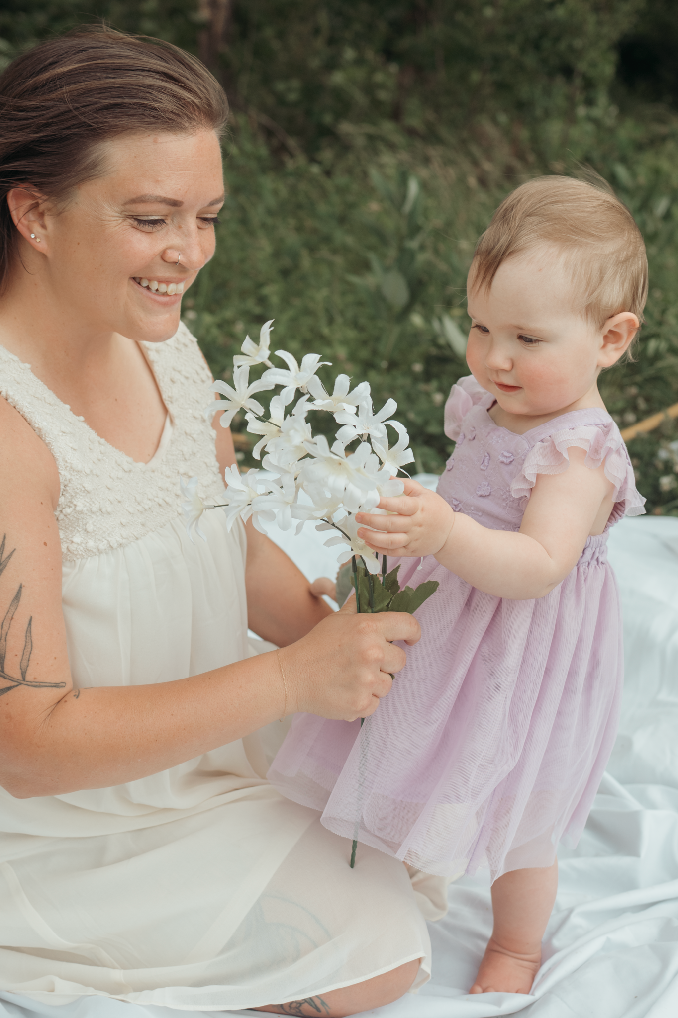 A mother and baby share a tender moment with white flowers while sitting on a blanket outdoors.