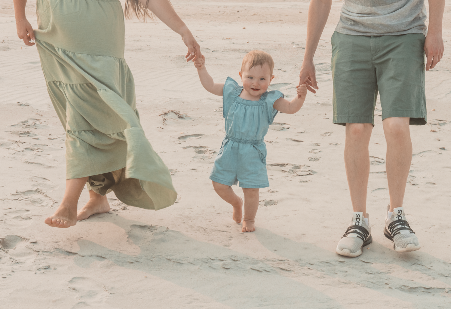 A small child in a blue outfit walks on the beach while holding hands with adults in casual beachwear.