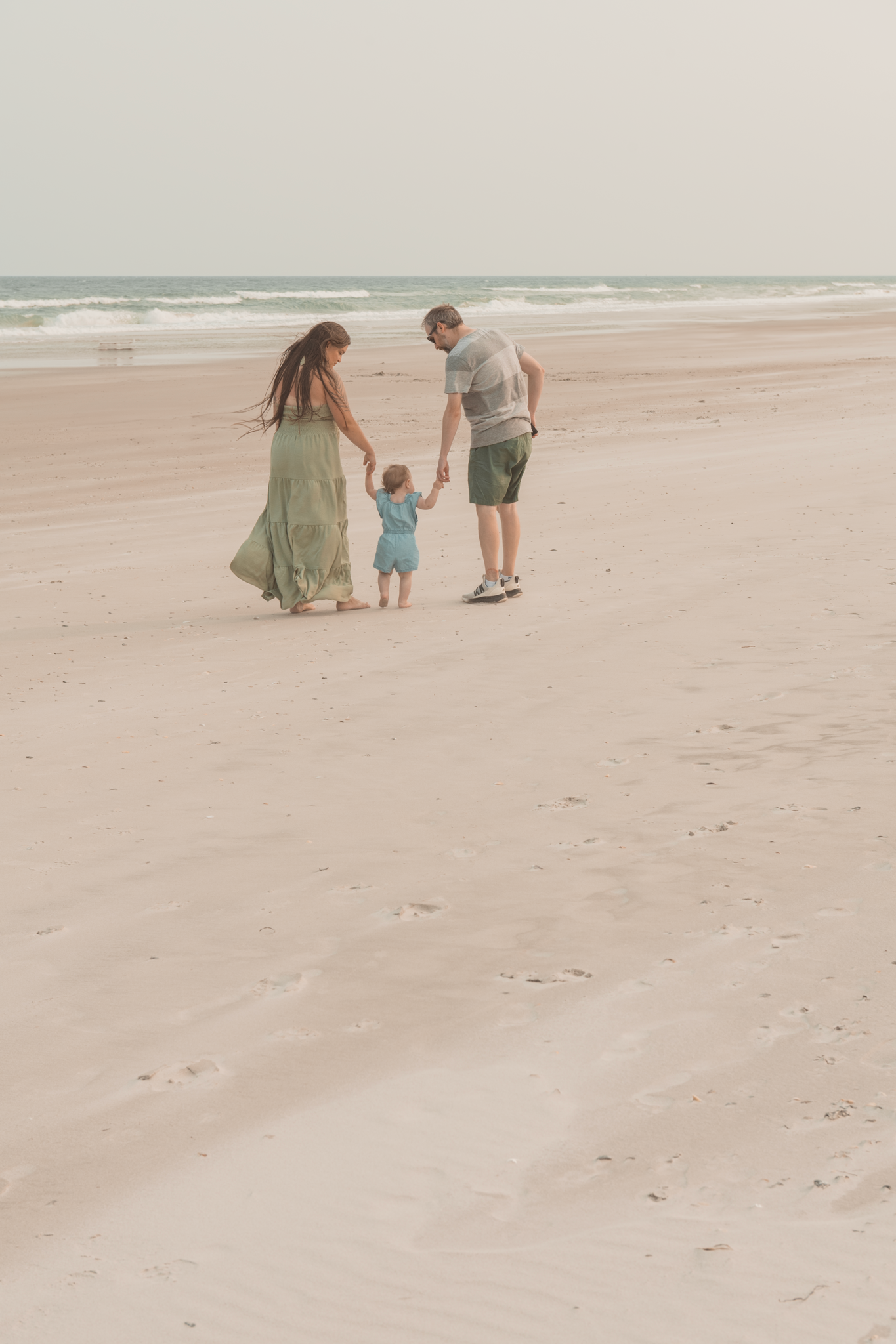 A couple and their small child walk hand in hand along a peaceful beach at sunset with waves rolling in the distance.