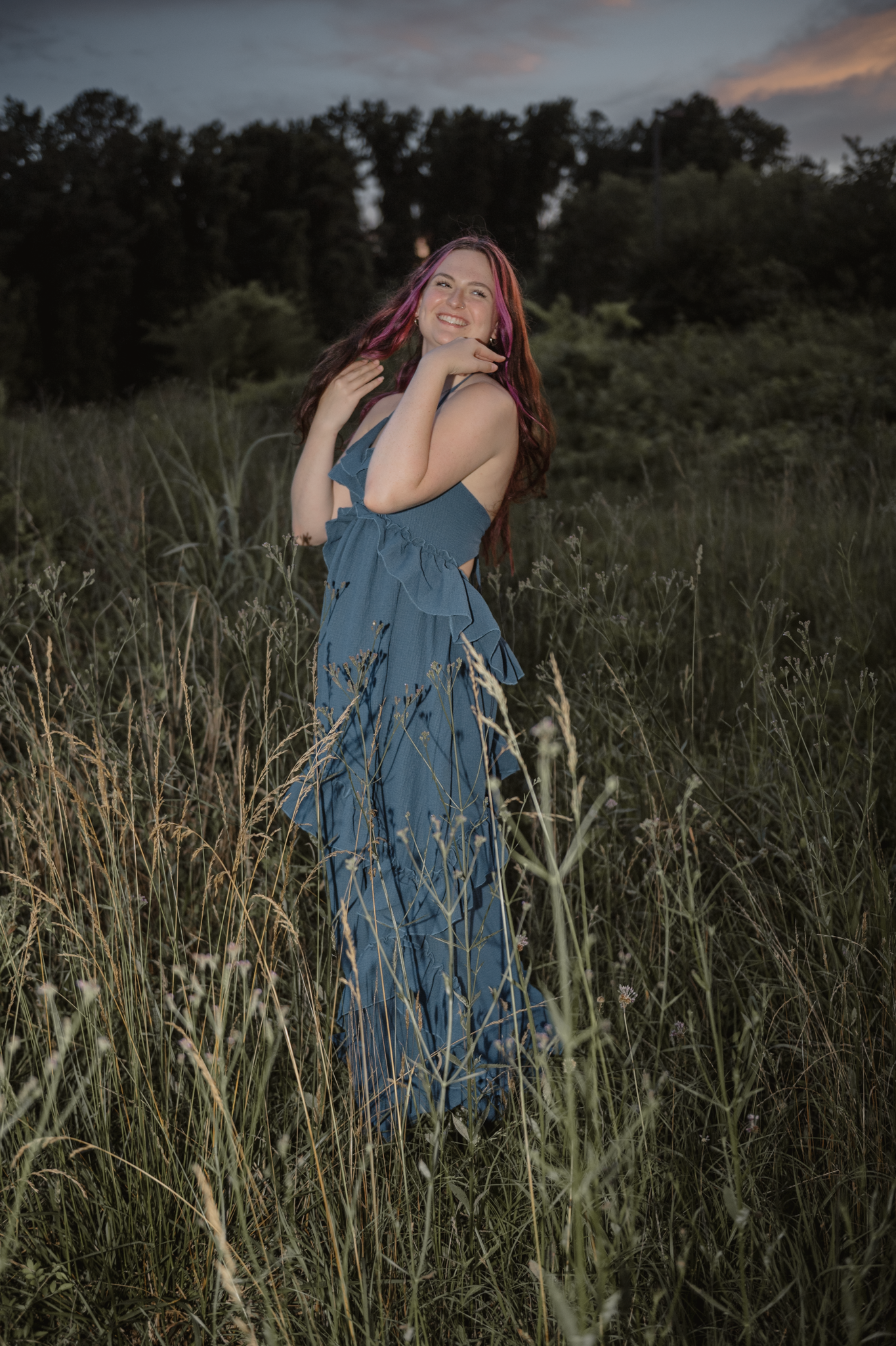 A long blue dress flows gracefully among tall grass at sunset in a dreamy outdoor portrait.