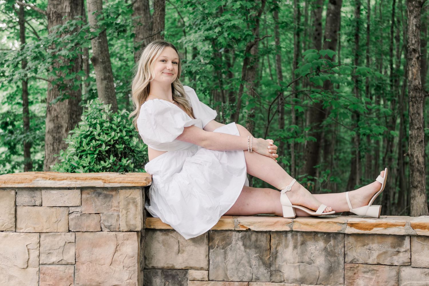 Someone in a white lace dress sits on a stone wall surrounded by lush green forest scenery.