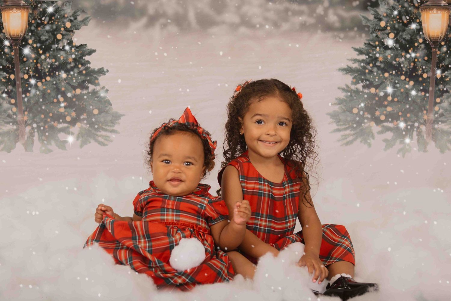 Two small children in matching red plaid holiday outfits sit together on a snowy backdrop with Christmas trees.