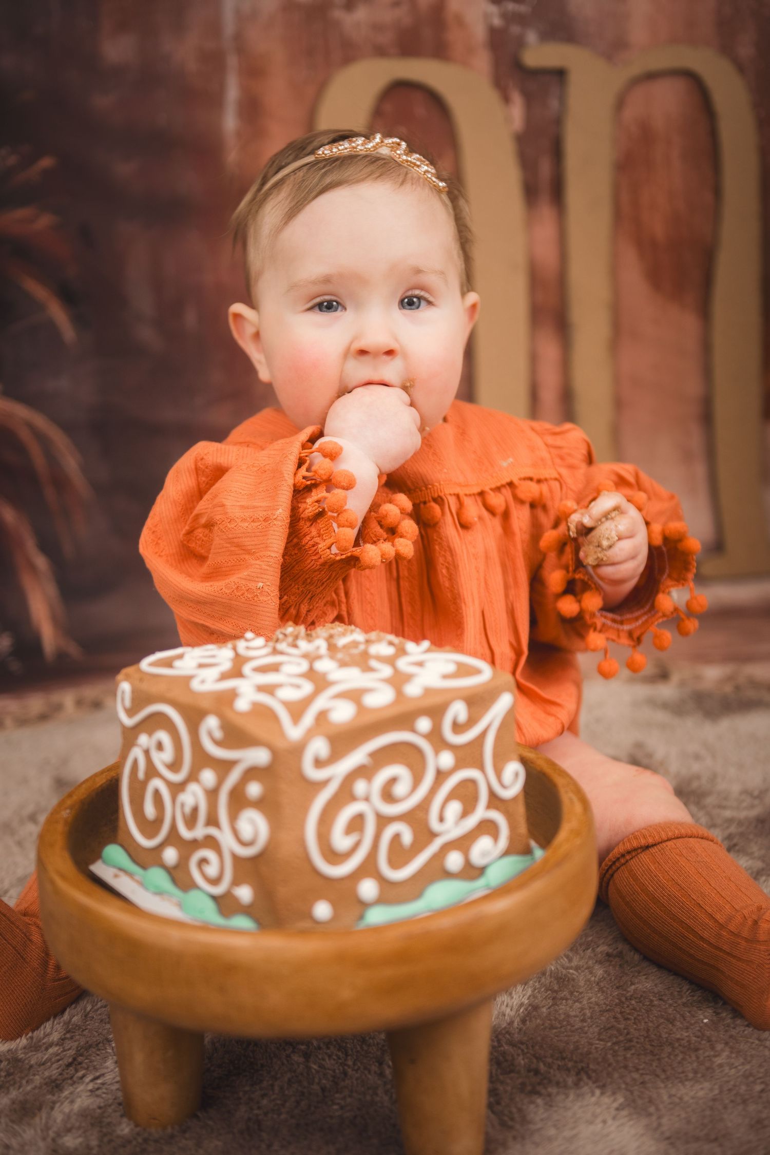 A small toddler in an orange dress sits next to a beautifully decorated chocolate cake on a wooden pedestal smashing her birthday cake in NC