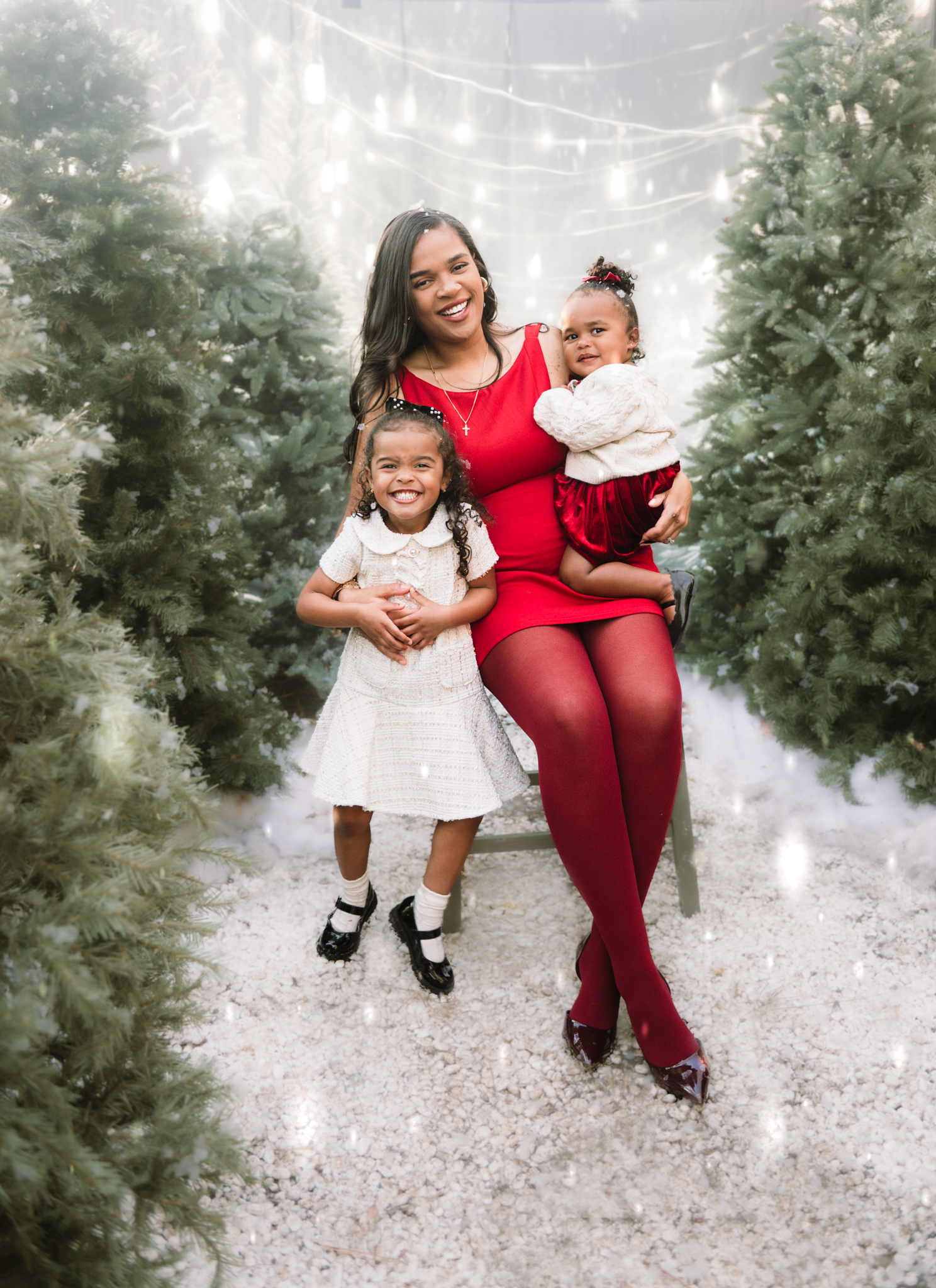 A festive holiday portrait in a snowy evergreen setting with string lights and coordinated red and white outfits.