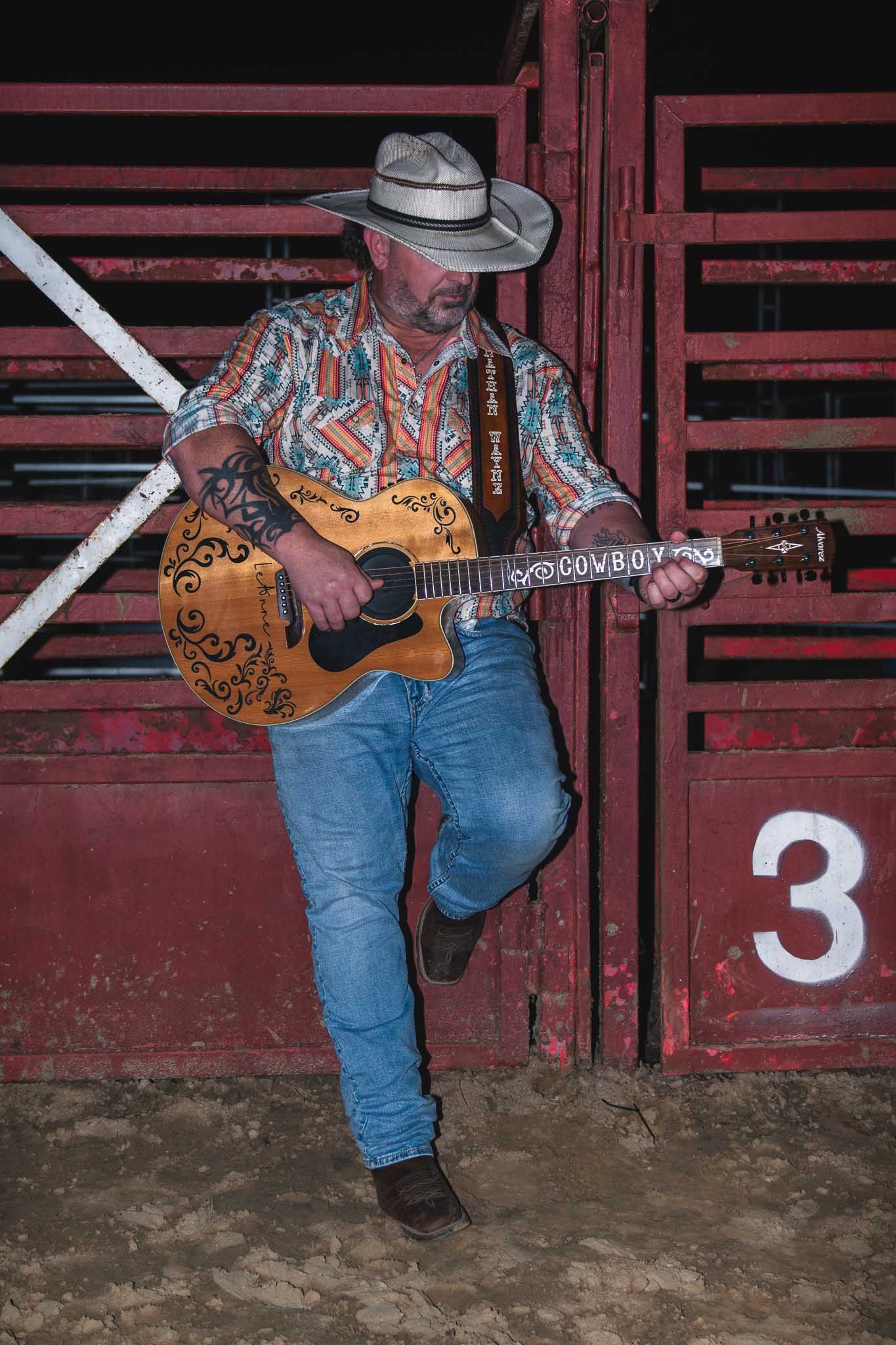 A musician in western attire sits on a wooden bench playing an acoustic guitar against a rustic red barn backdrop.