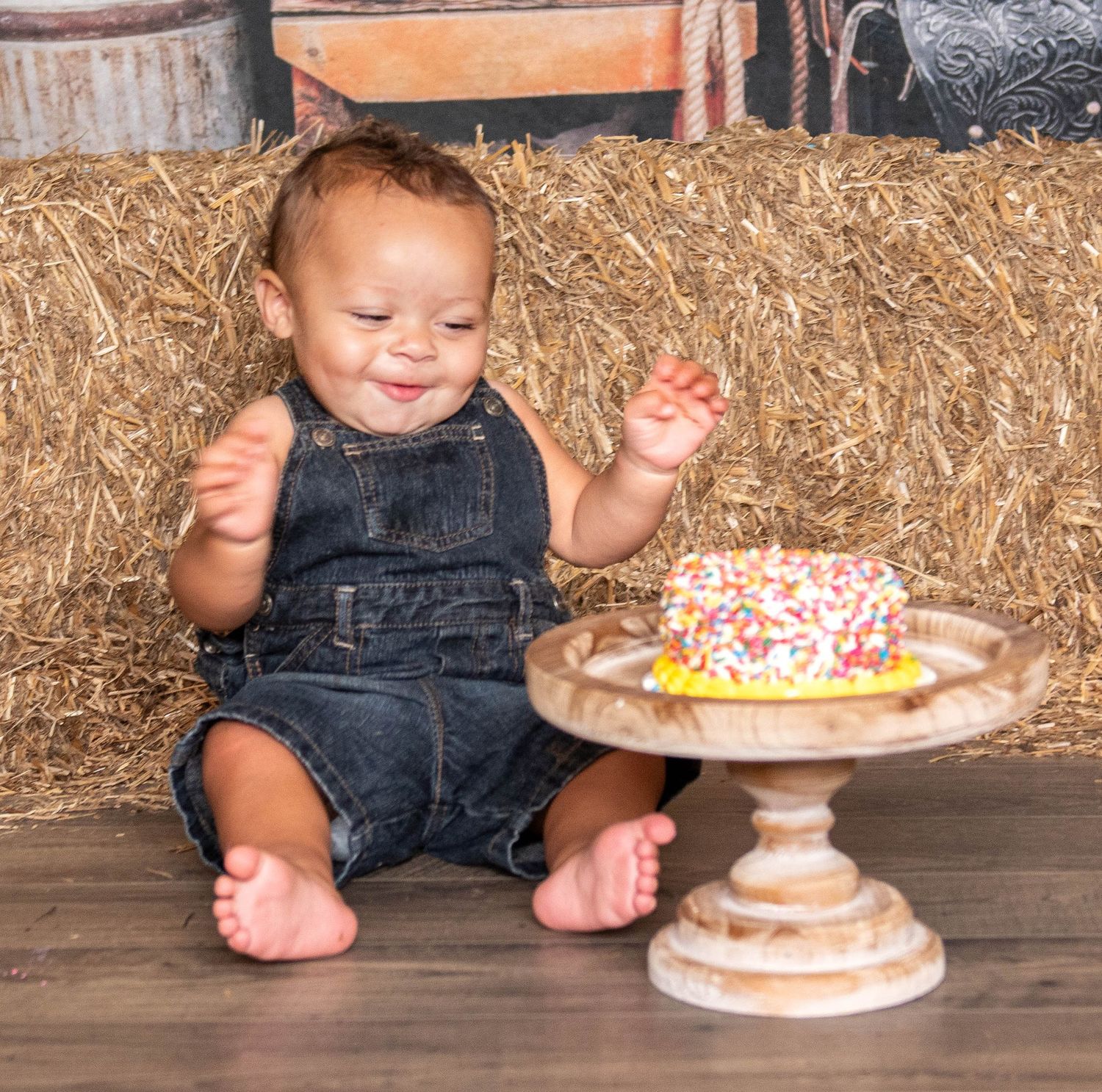 Toddler in denim overalls sits by hay bales with a decorated cake on a pedestal stand nearby.