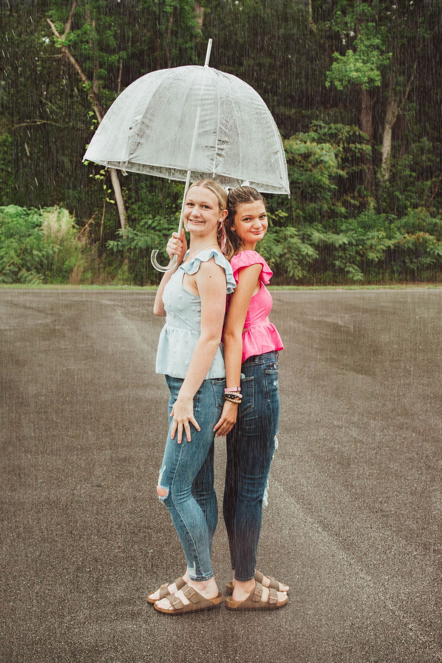 Two people stand back-to-back under a white umbrella in the rain, wearing casual clothing on a wet paved surface.