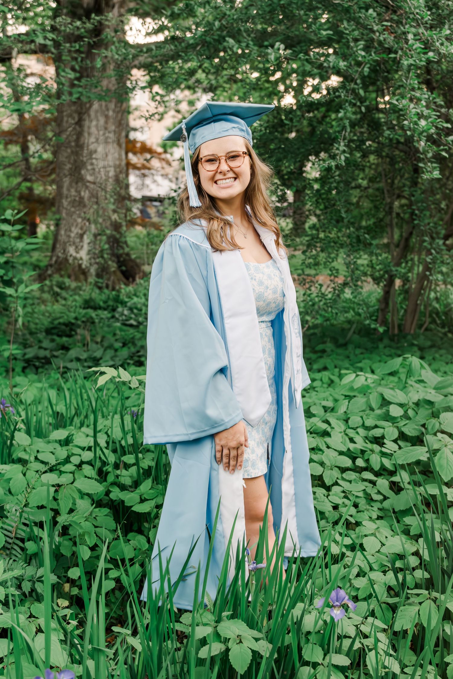 A graduate in light blue cap and gown poses outdoors among green foliage and flowers in a garden setting.