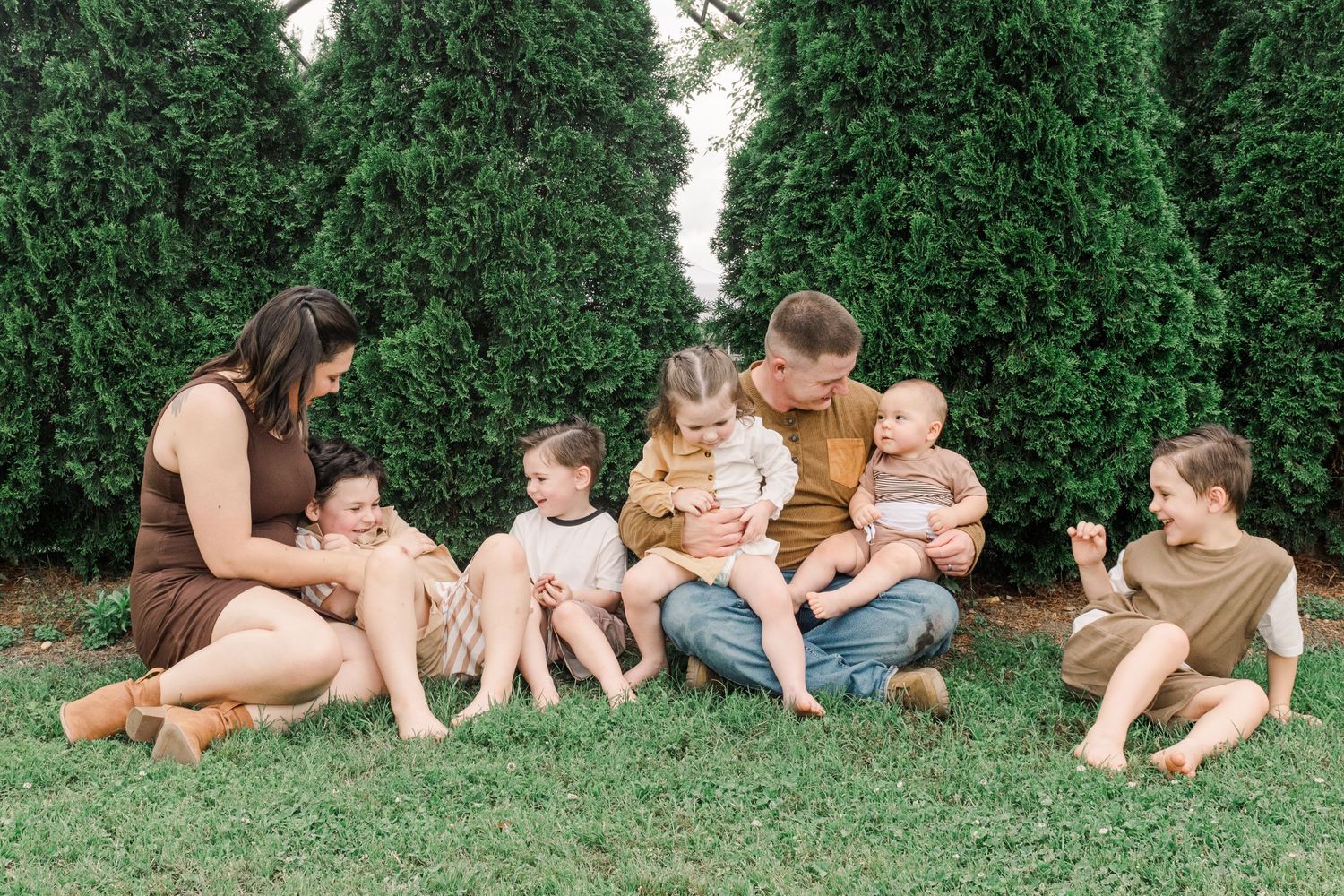 A family sits together on grass in front of a row of evergreen trees on a sunny day.