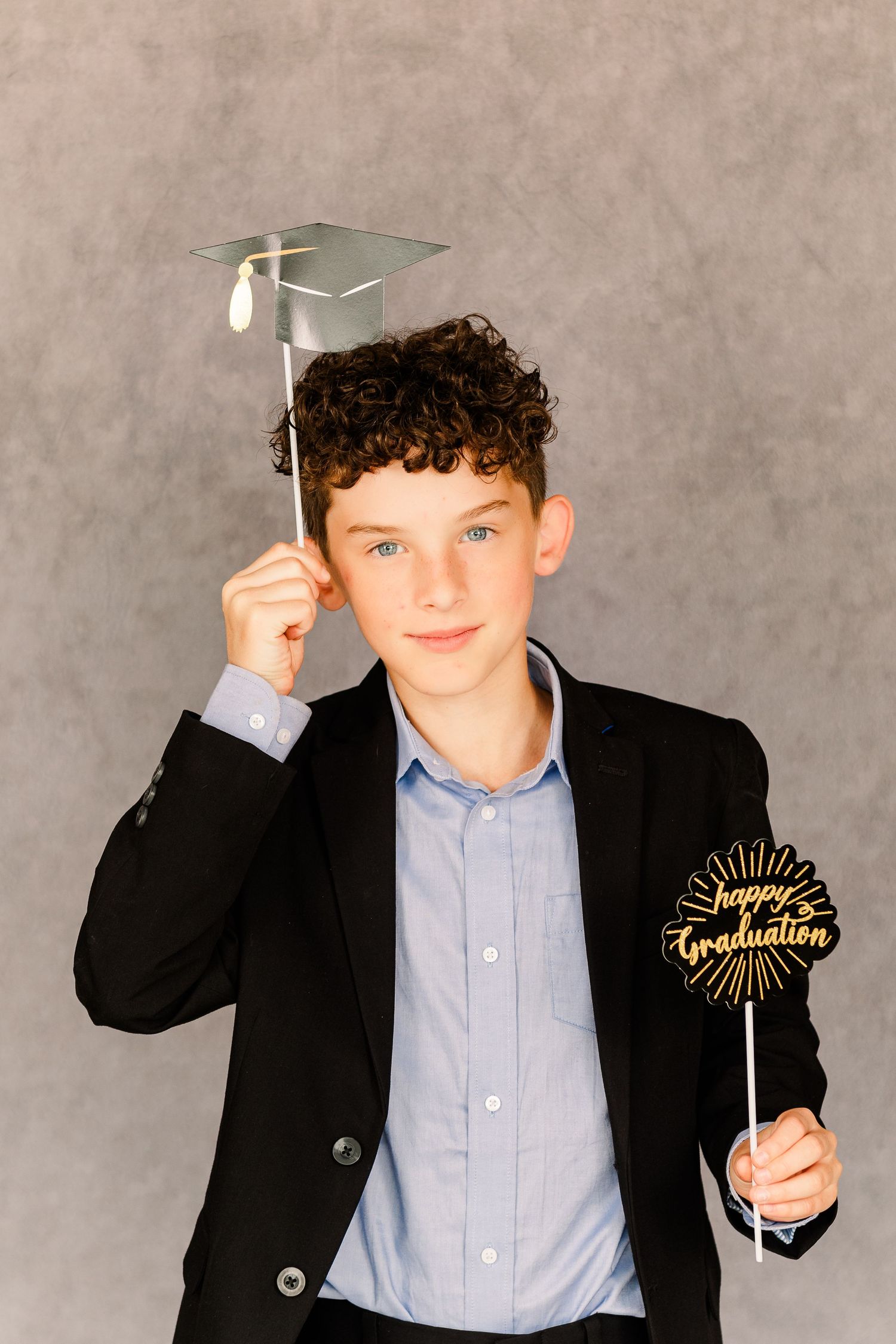 A graduate in a dark blazer and blue shirt holds props including a small graduation cap and decorative sign.