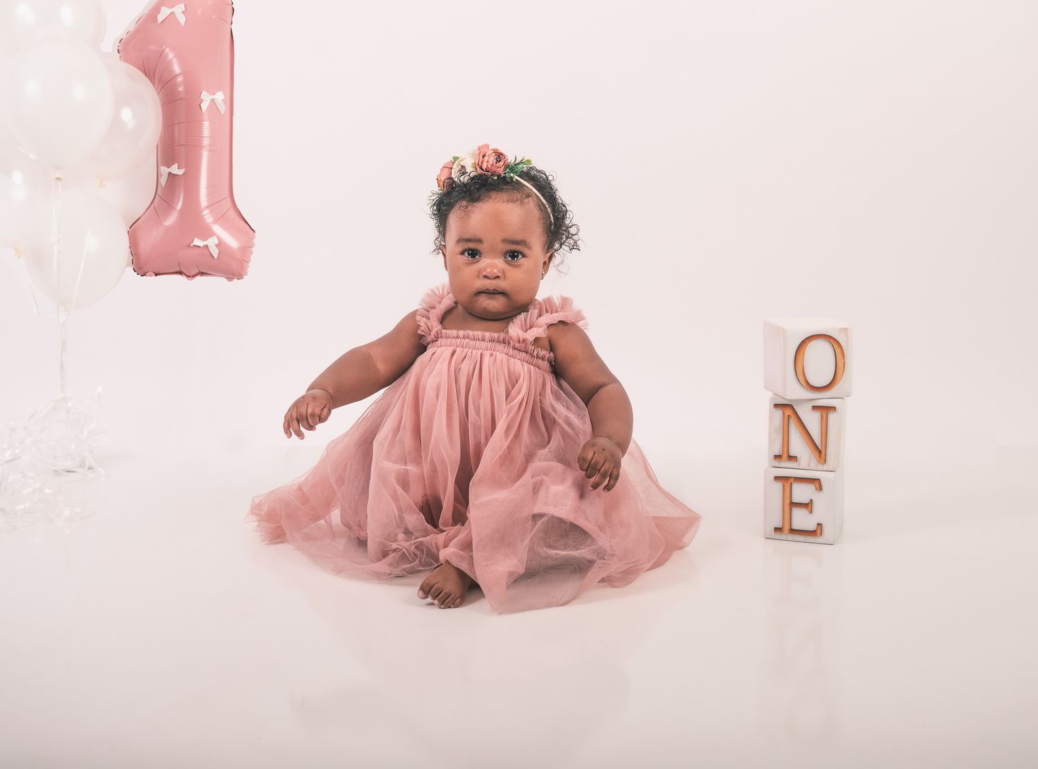 Baby in pink tulle dress sits on white backdrop with number one decoration and 'ONE' blocks for first birthday photo shoot.