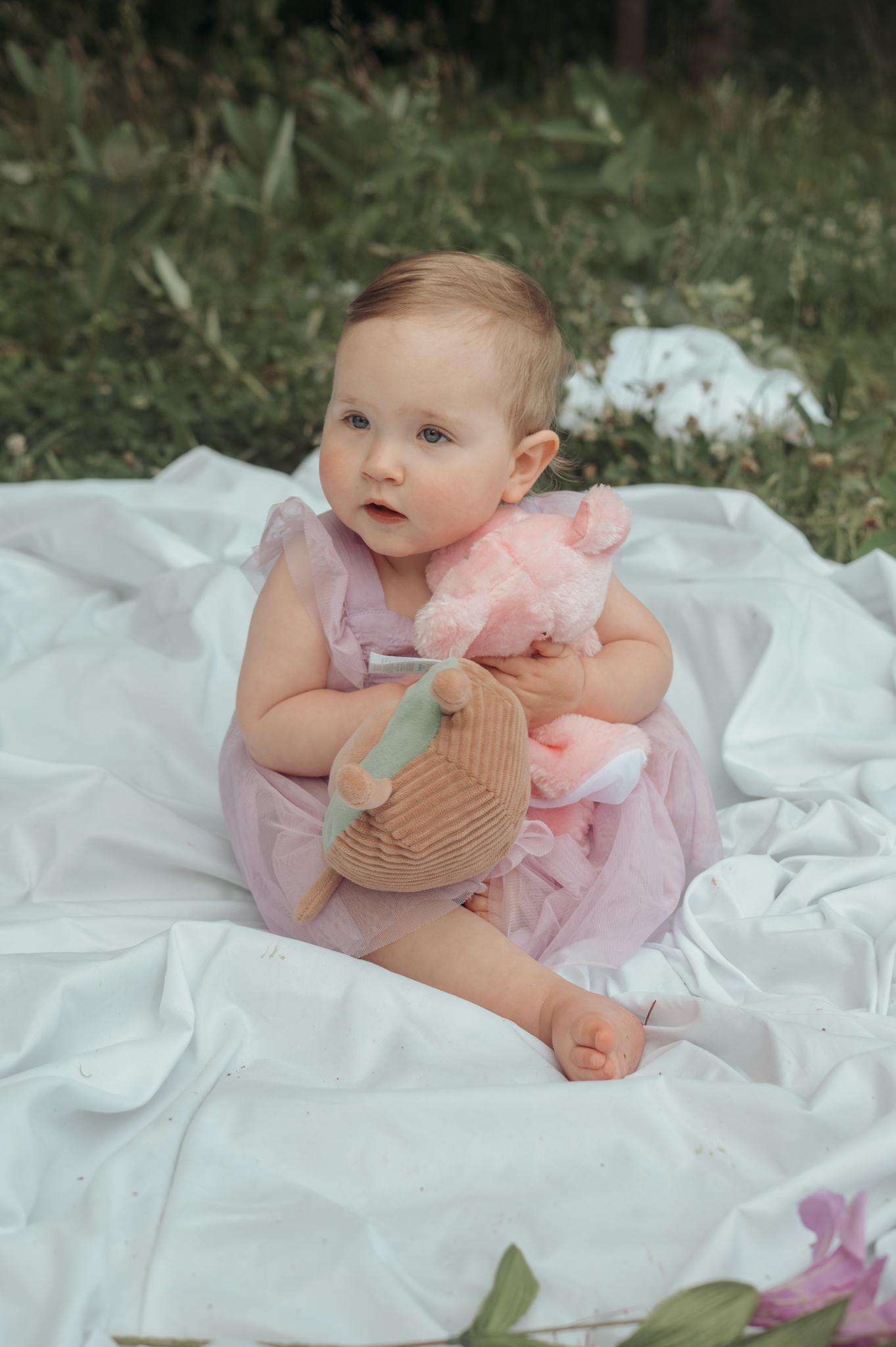 Small baby in pink dress sits on white blanket outdoors while holding a plush toy.