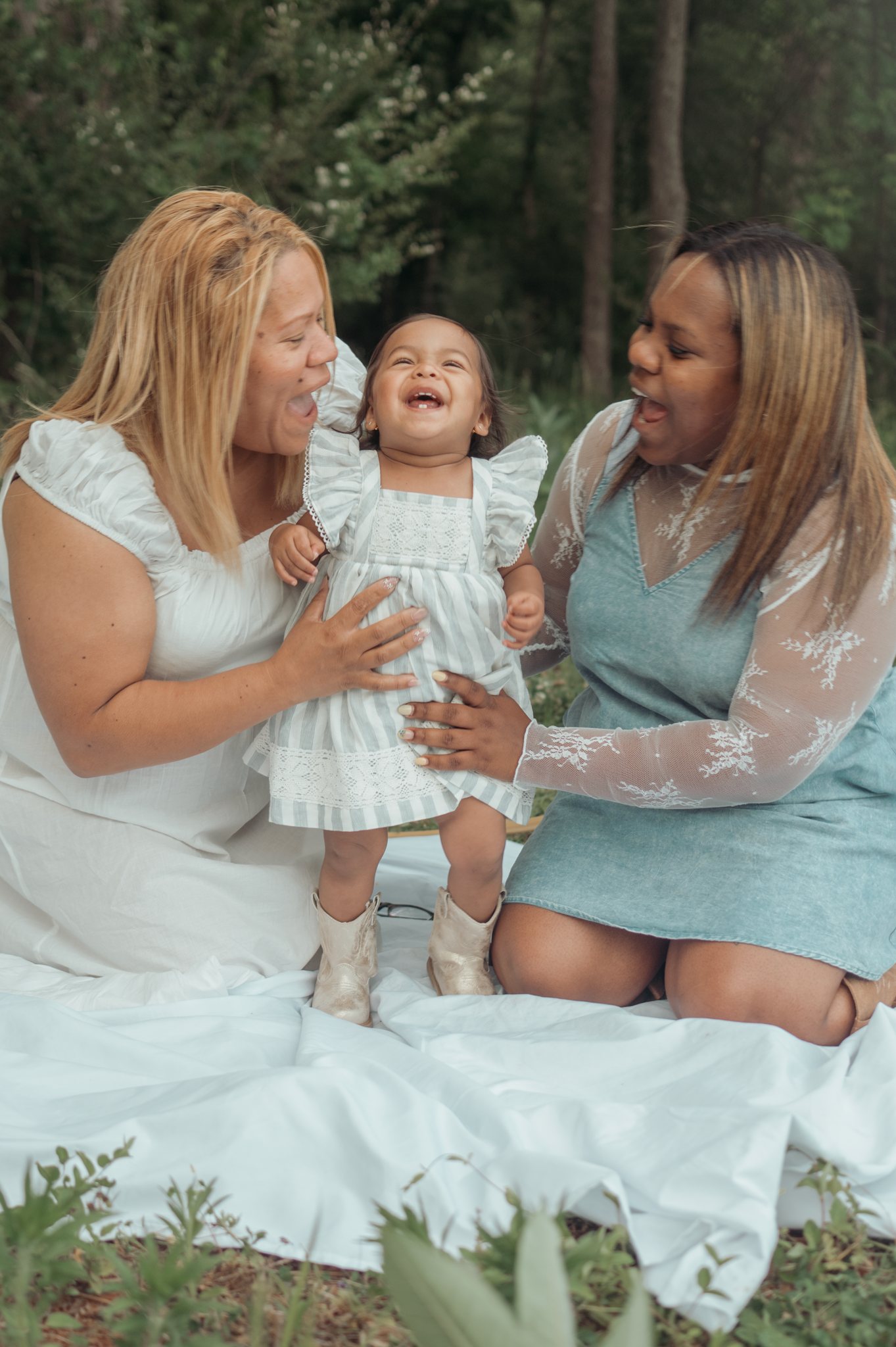 Three people laughing together outdoors while sitting on a white blanket in nature during a joyful photoshoot.
