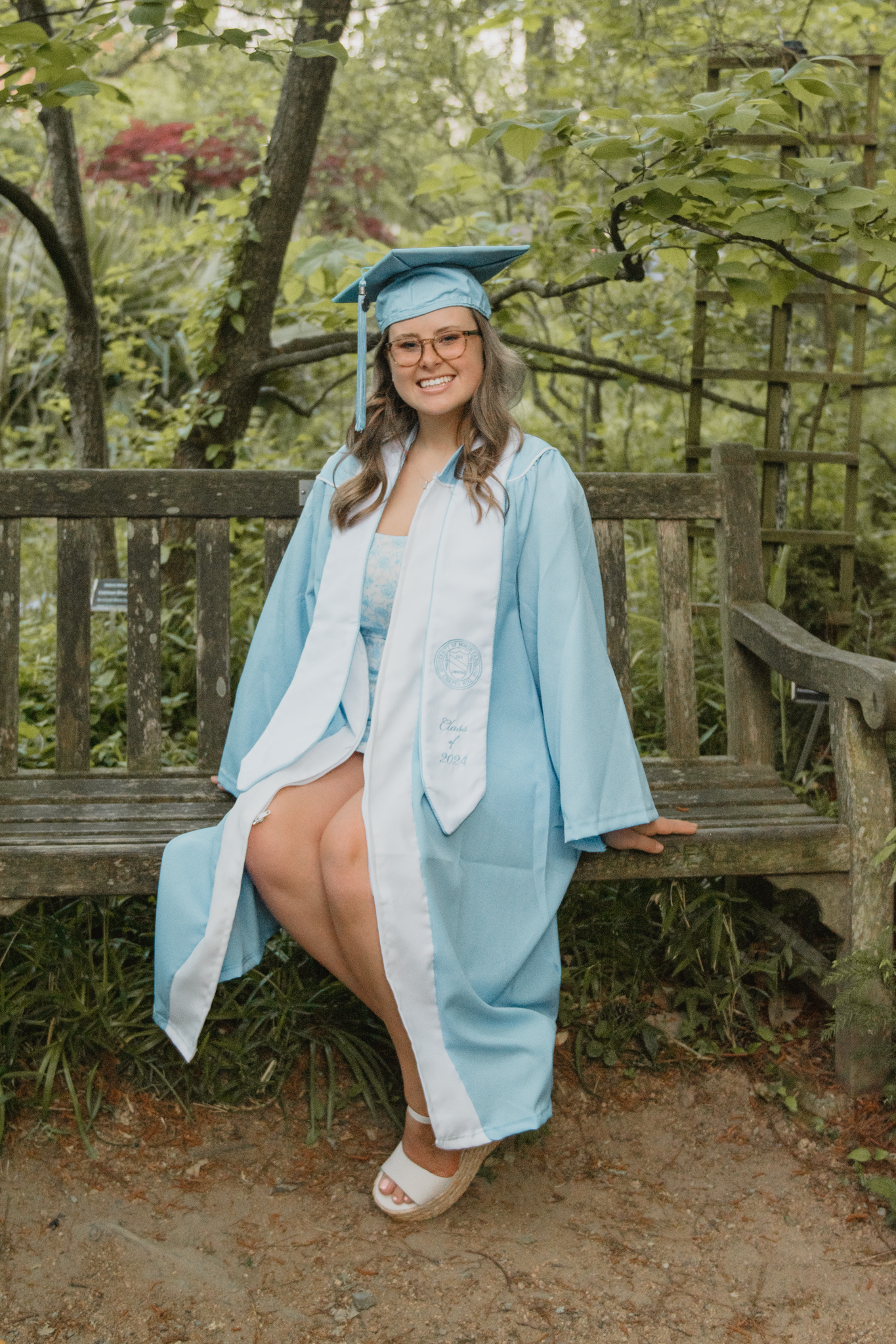 Graduate in light blue cap and gown sits on wooden bench in lush garden setting during graduation photoshoot.