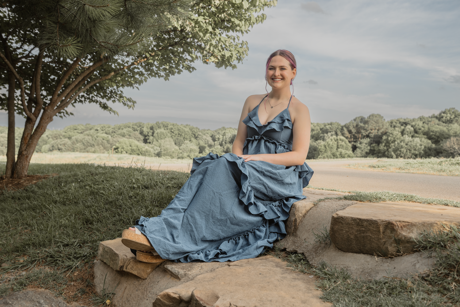 Haylee in a flowy blue dress sits on a stone ledge under a tree in an outdoor rural setting in Raleigh during her senior session
