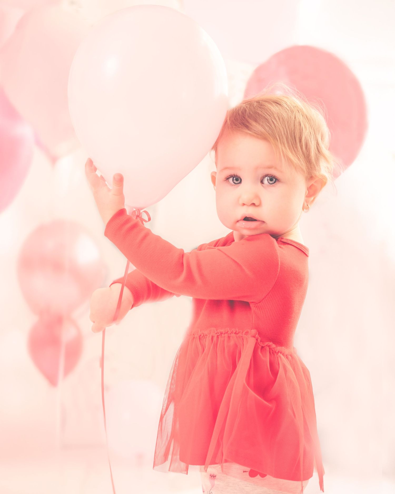 A small child in a red dress plays with pink balloons against a soft pastel background.